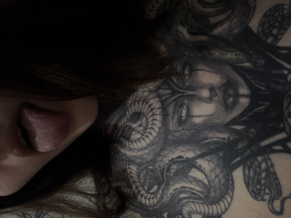 The image shows a person lying down with their tongue partially out of their mouth. The person has dark hair. A large, detailed tattoo covers part of their shoulder and upper back. The tattoo depicts a woman's face surrounded by snakes, resembling Medusa. The tattoo is black and gray with intricate shading.