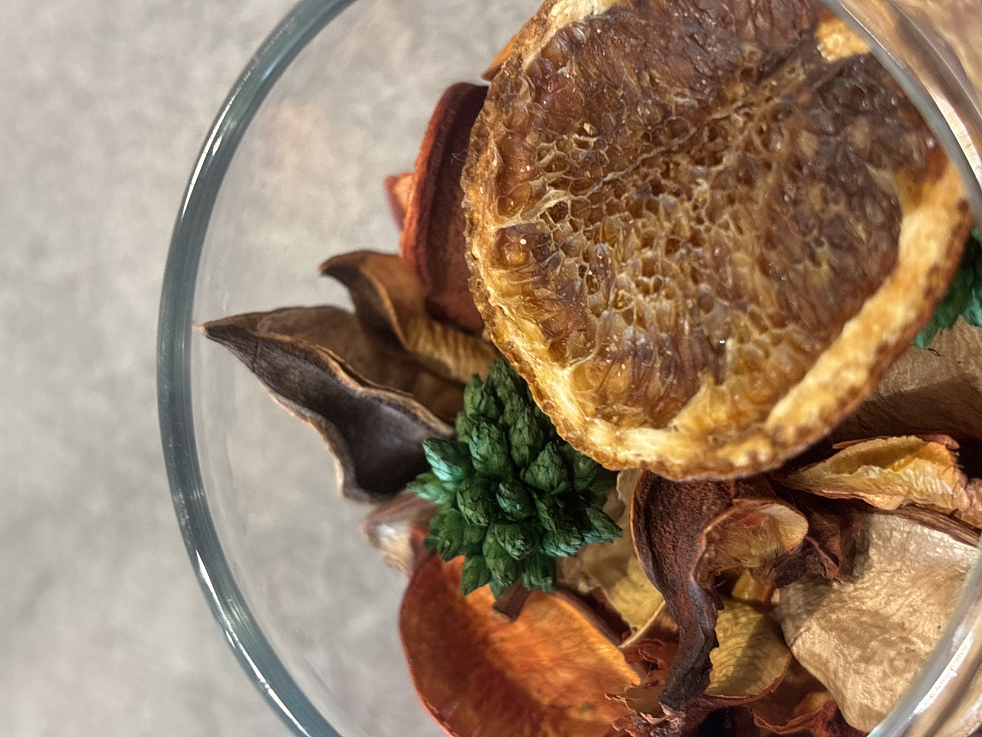 A close-up, high-angle shot shows a glass bowl containing potpourri. The potpourri consists of dried orange slices, various dried petals in shades of orange, brown, and beige, and a green, spiky, artificial decorative element. The bowl's clear glass is visible along the left edge. The background is a neutral, light gray surface. The location is not discernable from the image and therefore cannot be identified within Burgas, Bulgaria.