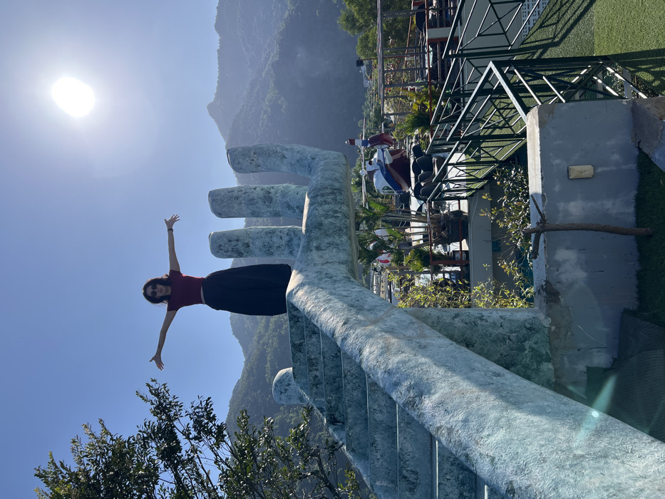 A female individual stands atop a large, light blue-grey concrete structure sculpted into the shape of a human hand, featuring integrated steps leading to the palm. She is positioned with arms outstretched and palms open, wearing a dark red crop top, a long dark skirt, and sunglasses, facing slightly left of the camera.
This scene is located at the "Hands of God" or "Golden Hand" attraction within VinWonders Nha Trang, situated on Hòn Tre Island, Nha Trang, Vietnam. The background displays a clear blue sky with a prominent sun and lens flare in the upper right, above a distant range of lush green, tree-covered mountains. Below the hand structure, the foreground reveals a multi-level outdoor area of the resort complex, containing a white, red, and blue helicopter display model, numerous potted plants, and black metal railings delineating pedestrian walkways. A large green-leafed tree partially frames the image on the far left. The environment is illuminated by direct, bright daylight.