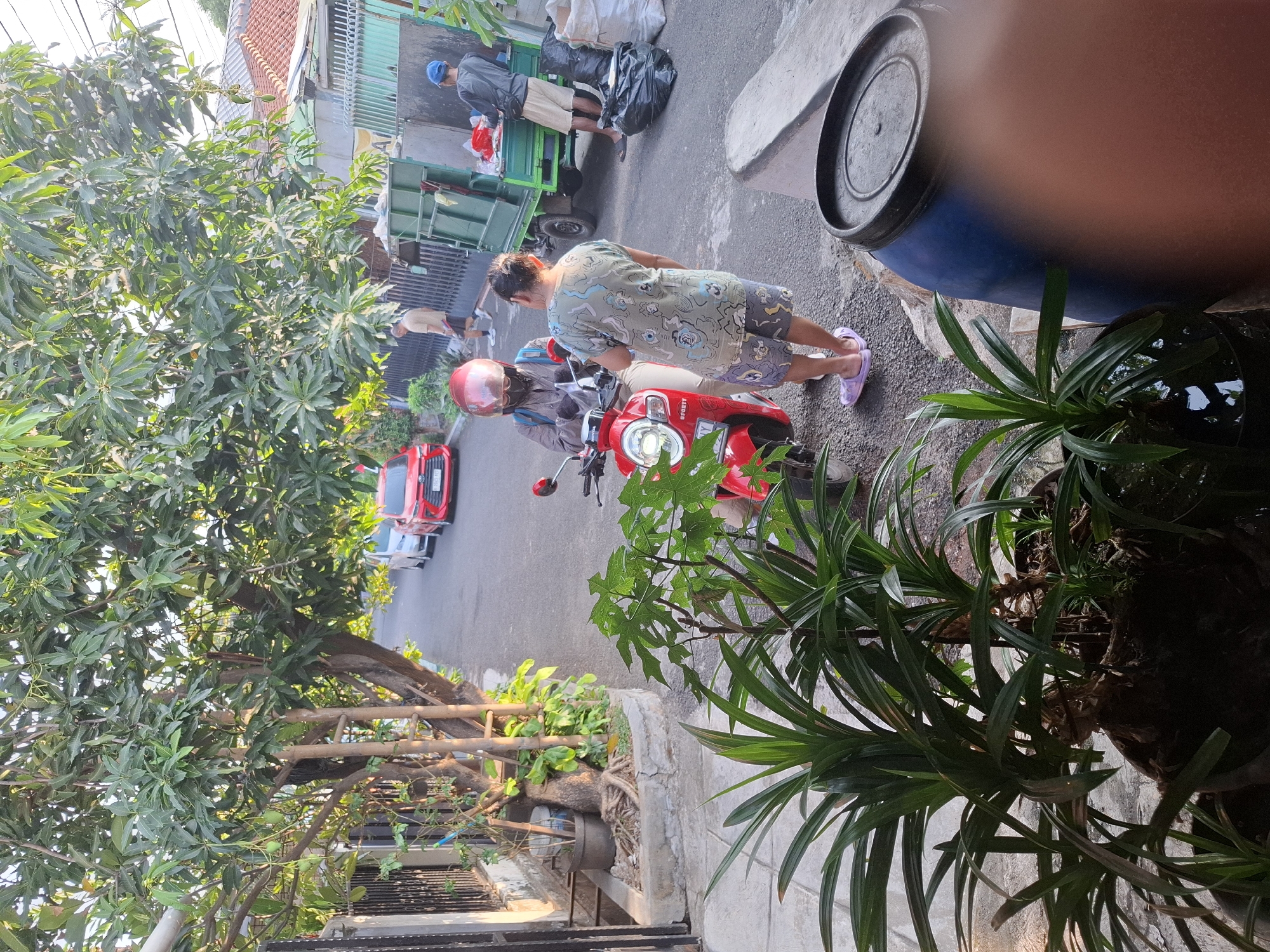 The image depicts a street scene in West Jakarta, Indonesia. In the foreground, several potted plants with long, green leaves are visible. To the right, a large blue barrel with a black lid is positioned near a concrete structure. In the middle ground, a red scooter is parked, with a person wearing a helmet and mask sitting on it. Another person, wearing a light-colored patterned shirt and patterned shorts, stands next to the scooter, facing the person on the scooter. Behind them, a green garbage truck is parked, with two individuals attending to it. One person, wearing a blue cap and shorts, is leaning into the truck. Another person, wearing a light-colored shirt and shorts, is walking away from the truck. Several plastic bags and trash are scattered around the truck. The street is paved, and there are trees and houses lining the street. A red car is parked further down the street. The sun appears to be setting, casting long shadows and creating a warm, golden light.