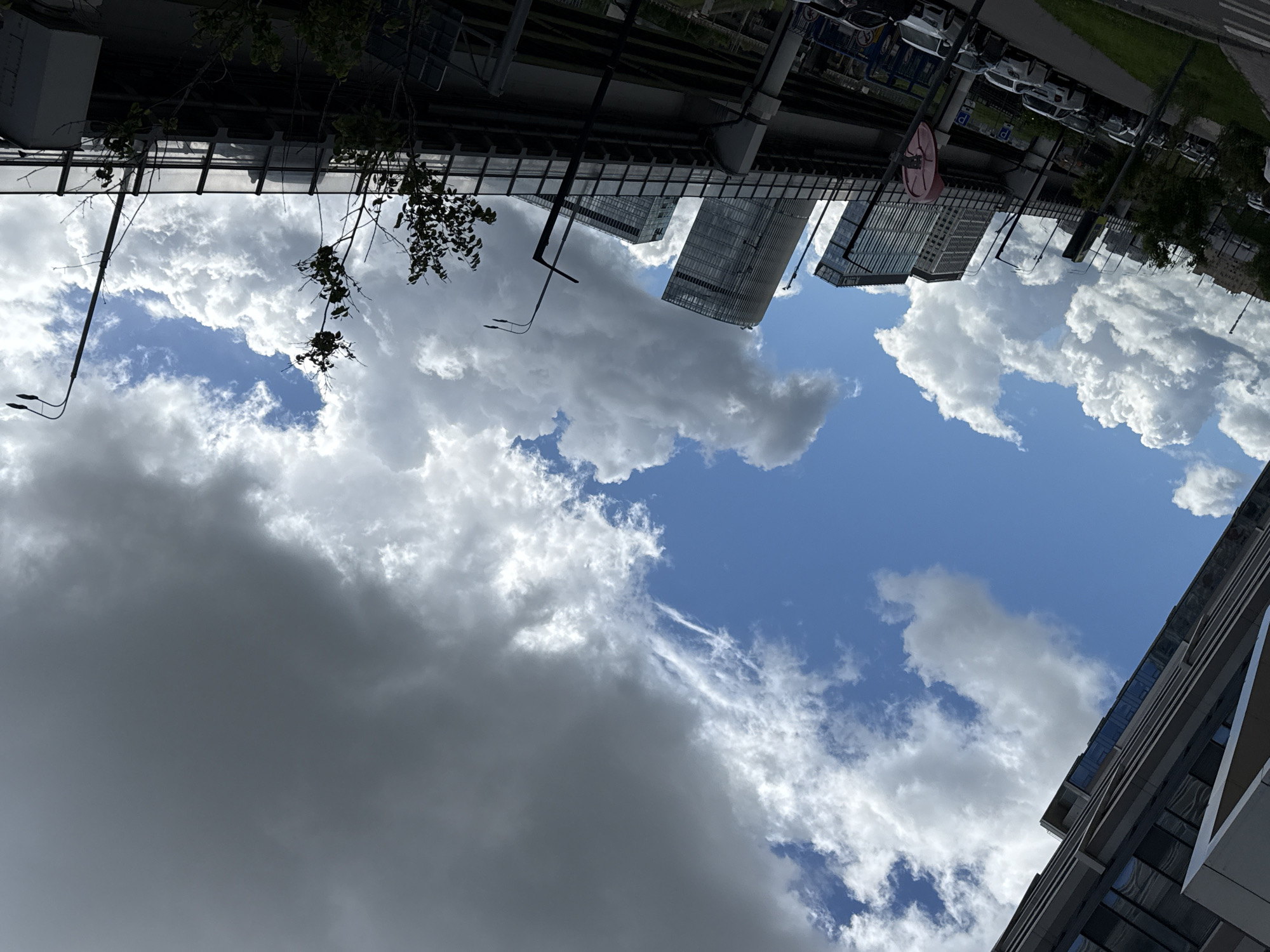 The image is taken from a low angle, looking up at the sky with buildings framing the shot. The sky is a mix of blue and white, fluffy clouds. In the upper portion of the frame, the bottom of a bridge or elevated structure is visible, along with some cars parked along the side of the road and street signs. The architecture suggests an urban environment, with modern skyscrapers in the background. Based on the overall style and context clues (skyscrapers, street signs, architecture) it is in Moscow, Russia.