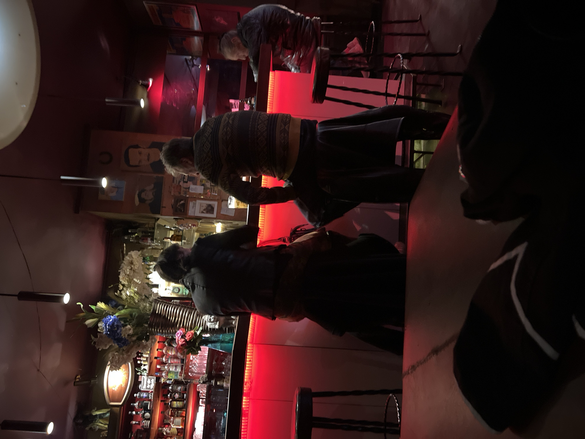 The image shows the interior of a bar in Berlin, Germany. The bar is illuminated with red lighting behind the counter. Several people are standing at the bar. One man is wearing a patterned sweater. Assorted liquor bottles are displayed on shelves behind the bar, along with floral arrangements. The bar counter is made of dark stone. A pendant light fixture is visible above the bar. The scene is dimly lit, creating a relaxed atmosphere.