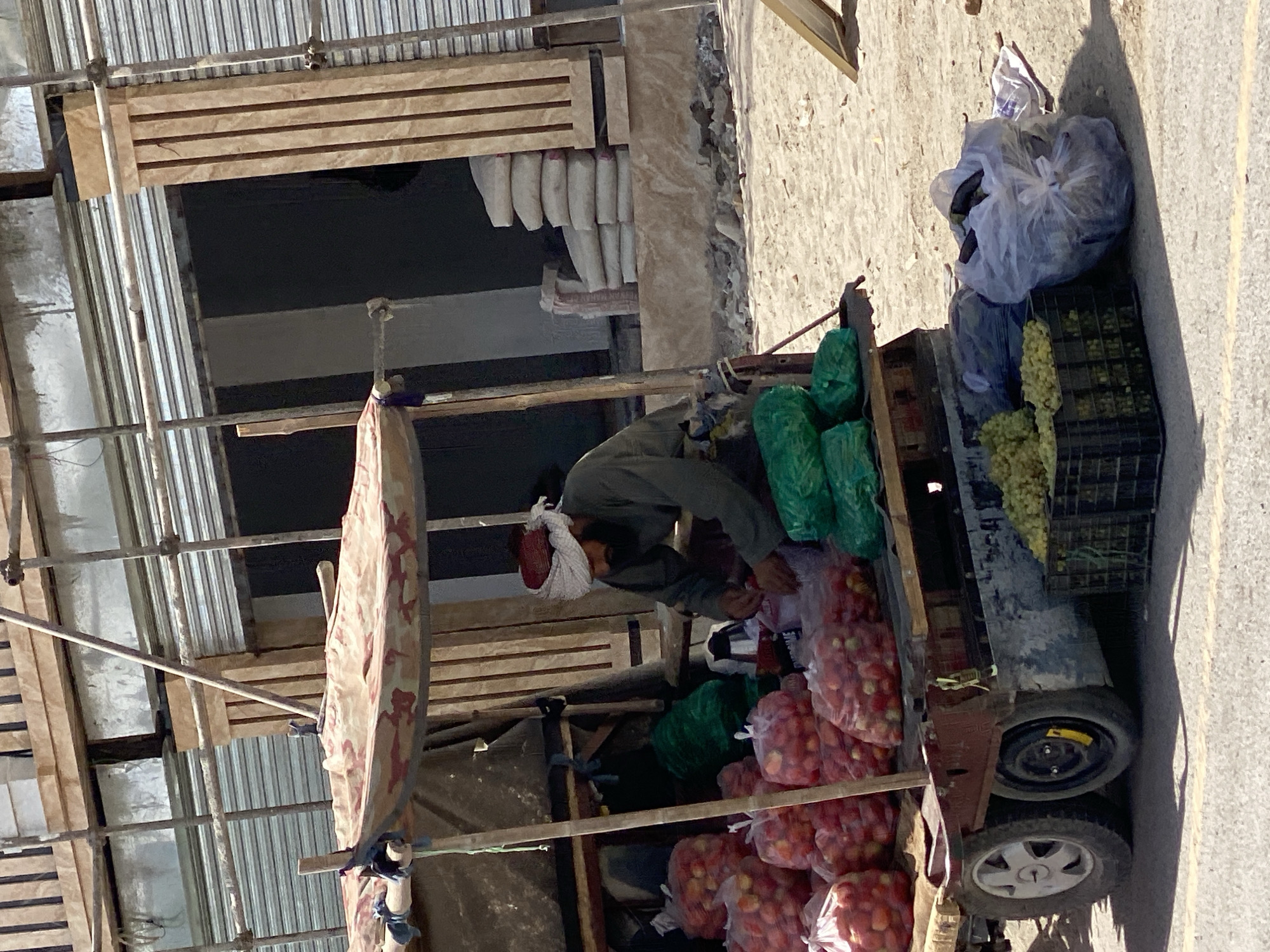 A man, wearing a turban and traditional clothing, is working at a fruit stall in Kandahar, Afghanistan. He is sitting behind a cart laden with bags of tomatoes and bunches of grapes. The cart is parked on a dirt road in front of a building under construction, with scaffolding and building materials visible. The man appears to be organizing or preparing the fruit for sale. The scene is set in a dusty, outdoor environment, typical of a marketplace.