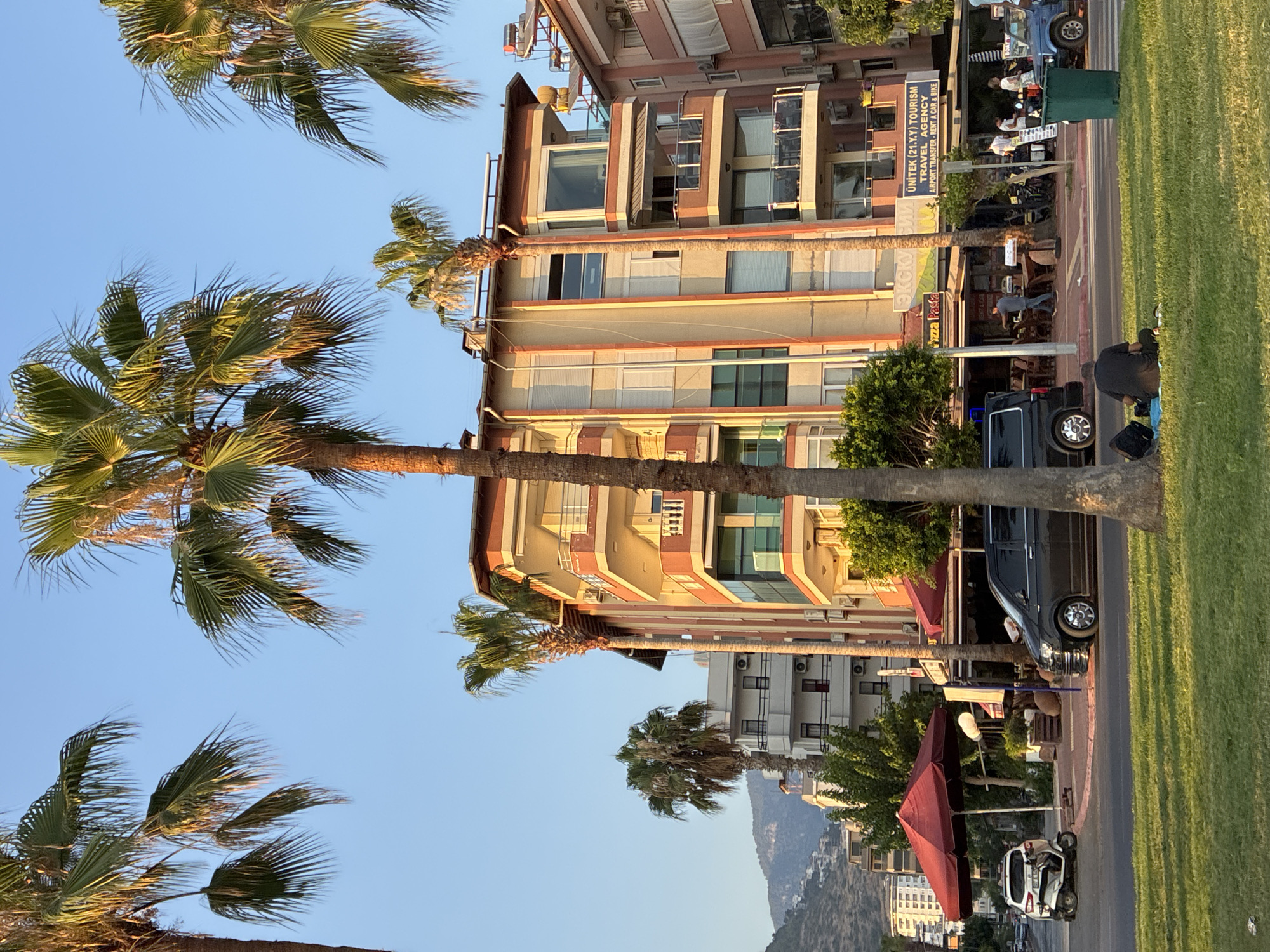 The image captures a street scene in Alanya, Turkey, during what appears to be late afternoon or early evening, given the warm lighting. The focus is on a multi-story building with balconies, painted in shades of peach and white, with red trim. Several palm trees are prominent in the foreground and framing the scene.
On the street level of the building, there are businesses, including a restaurant with outdoor seating and an umbrella providing shade. Signs indicate "Pizza Pasta" and "UNITЕК (21.Y.Y) TOURISM TRAVEL AGENCY AIRPORT TRANSFER - RENT A CAR & BIKE". A black van is parked on the right side of the street, and a white scooter is parked on the left.
In the grassy area in front of the street, a person is sitting on a blue mat, facing away from the camera, with a black backpack beside them. Other people are visible walking on the sidewalk near the businesses. In the background, more buildings and a mountain slope with scattered structures are visible under a clear blue sky. The scene suggests a typical urban environment in a coastal tourist destination.