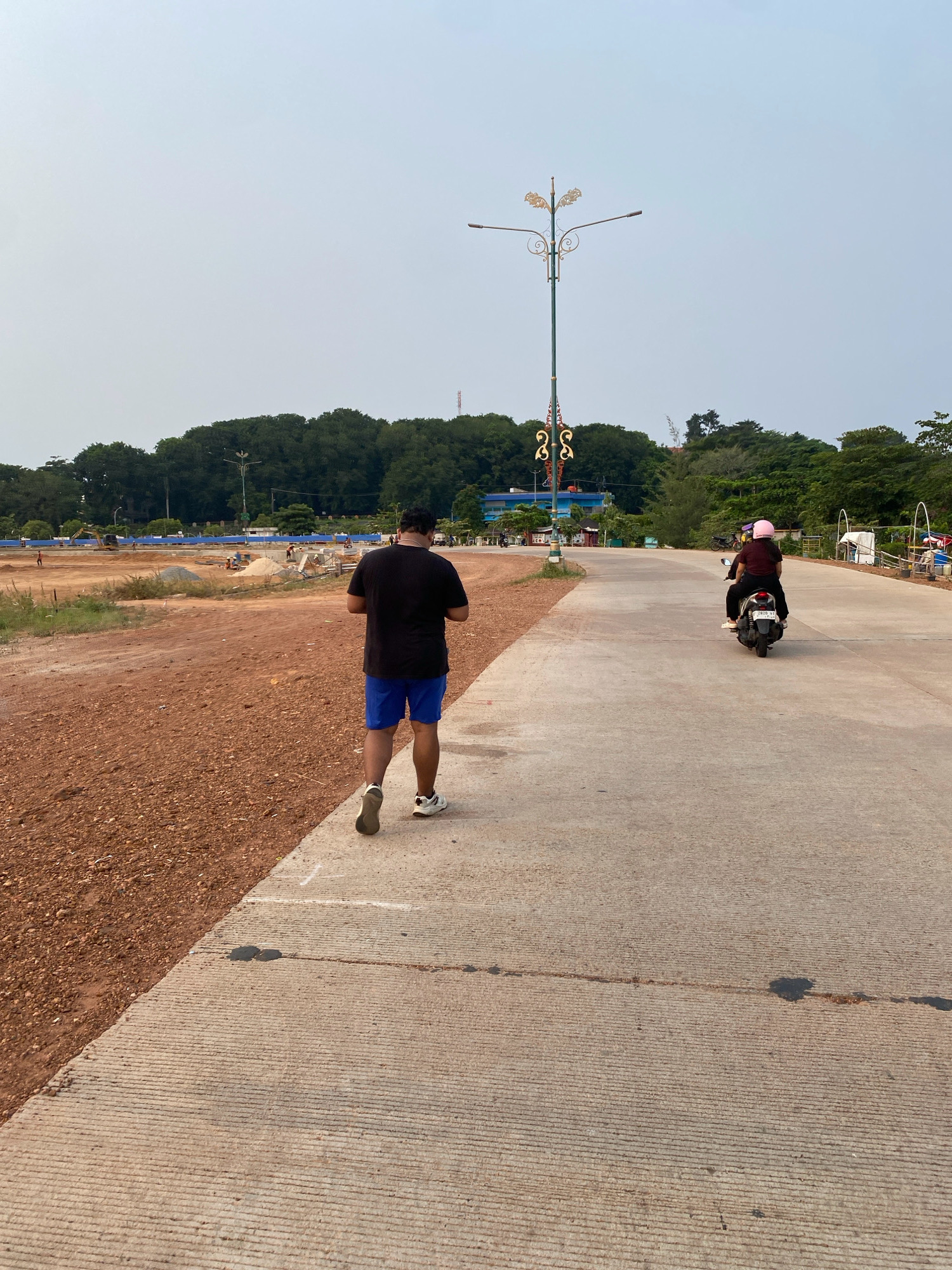 The image depicts an outdoor scene in Tanjung Pinang, Indonesia, featuring a concrete pathway next to a dirt construction area. A man in a black t-shirt and blue shorts is walking away from the camera on the pathway. Further down the pathway, a person wearing a pink helmet rides on the back of a motorcycle driven by another individual. In the background, a line of trees forms a backdrop with a distinctive decorative streetlamp visible. The construction area shows exposed red soil, some mounds of dirt, and visible construction equipment. The sky is overcast, suggesting a hazy or cloudy day. The location appears to be near a body of water or coastal area given the vegetation and open space.