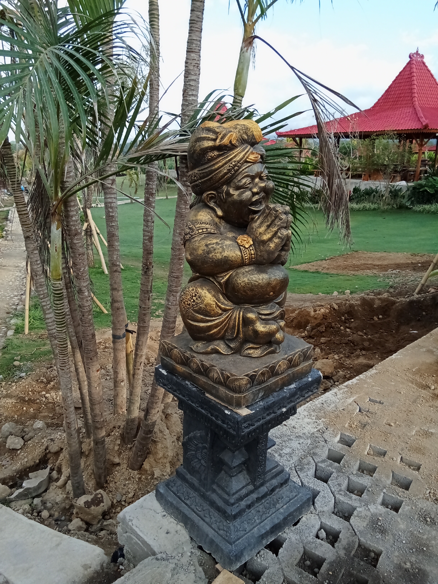 In Selemadeg Timur, Indonesia, an outdoor scene features a golden statue of a stylized human figure in a praying or pleading posture, adorned with a distinctive headdress and jewelry. The statue is mounted on a dark, intricately carved pedestal, which rests on a light-colored concrete base. Surrounding the statue are several palm trees with rough bark, some tied with dark bands. The ground is a mix of earth, gravel, and scattered rocks. In the background, a vibrant red-roofed gazebo with visible wooden supports stands amidst a manicured green lawn. The overall setting suggests an outdoor recreational or cultural space undergoing some landscaping or construction, indicated by exposed soil and partially laid paving stones.