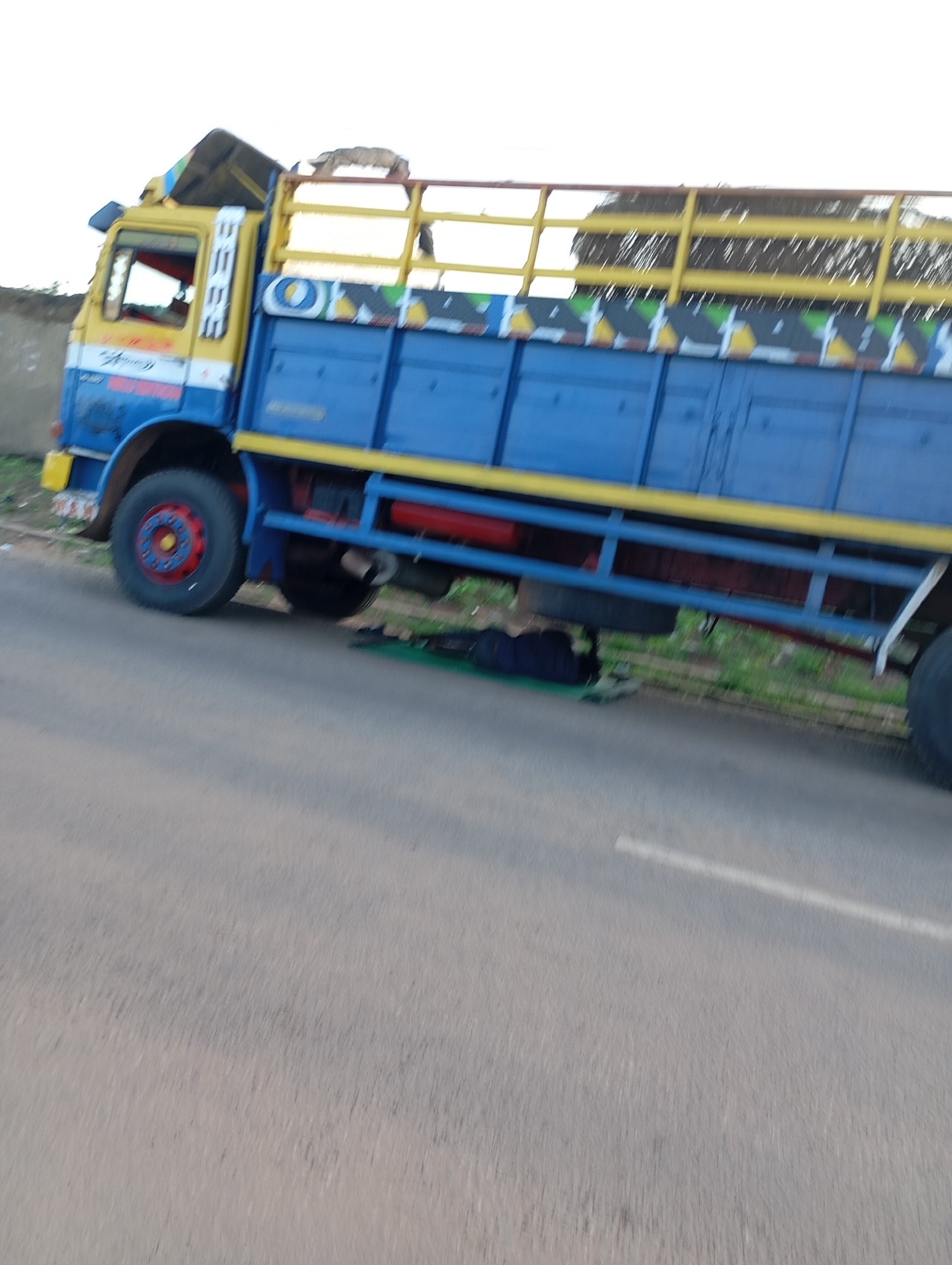 A yellow and blue truck with a damaged roof and yellow railings is parked on the side of a paved road. The truck has a blue cargo bed with yellow trim. A red fuel tank is visible beneath the cargo bed. The road has a white line painted on it. The environment appears to be grassy with some vegetation. A dark blue bag is placed on a green mat on the ground next to the truck.