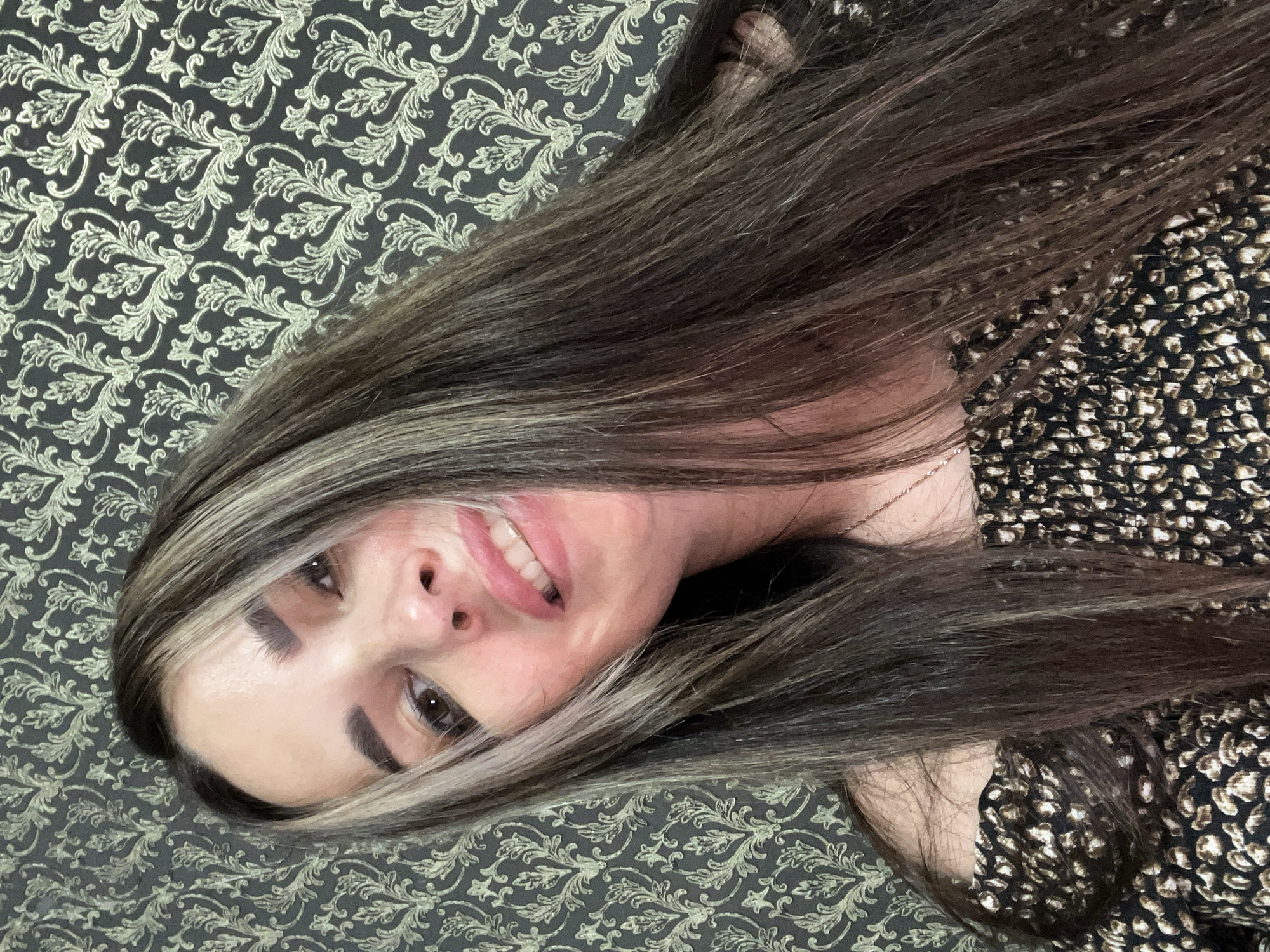 A woman with long, dark brown hair with blonde highlights, wearing an off-the-shoulder top with a dark background and a light brown animal print pattern, is captured in a selfie. She is looking directly at the camera with her mouth slightly open, revealing her teeth. The background is a dark wallpaper with a metallic gold floral pattern. The scene appears to be indoors, likely in a residential setting, given the wallpaper. The lighting is artificial, casting soft shadows on her face. The image is taken from a low angle, looking up at the woman.