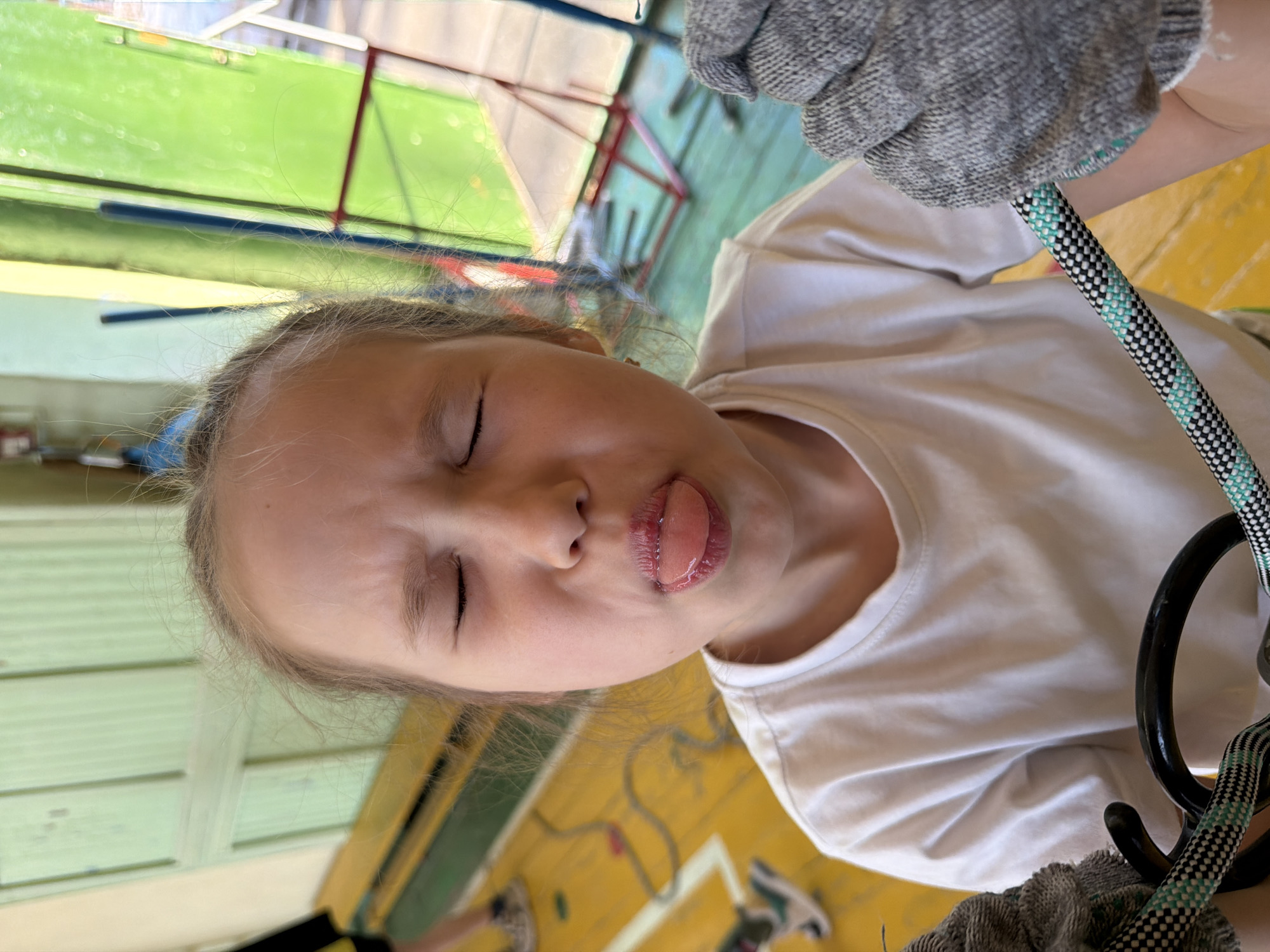 A photograph shows a young girl with fair skin, light brown hair, and closed eyes, sticking her tongue out. She is wearing a white t-shirt and gray gloves. A climbing rope and equipment are visible in the foreground, suggesting she is engaged in a climbing activity. The background reveals a gym or climbing facility with padded walls and other climbing structures. The location is Izhevsk, Russia.