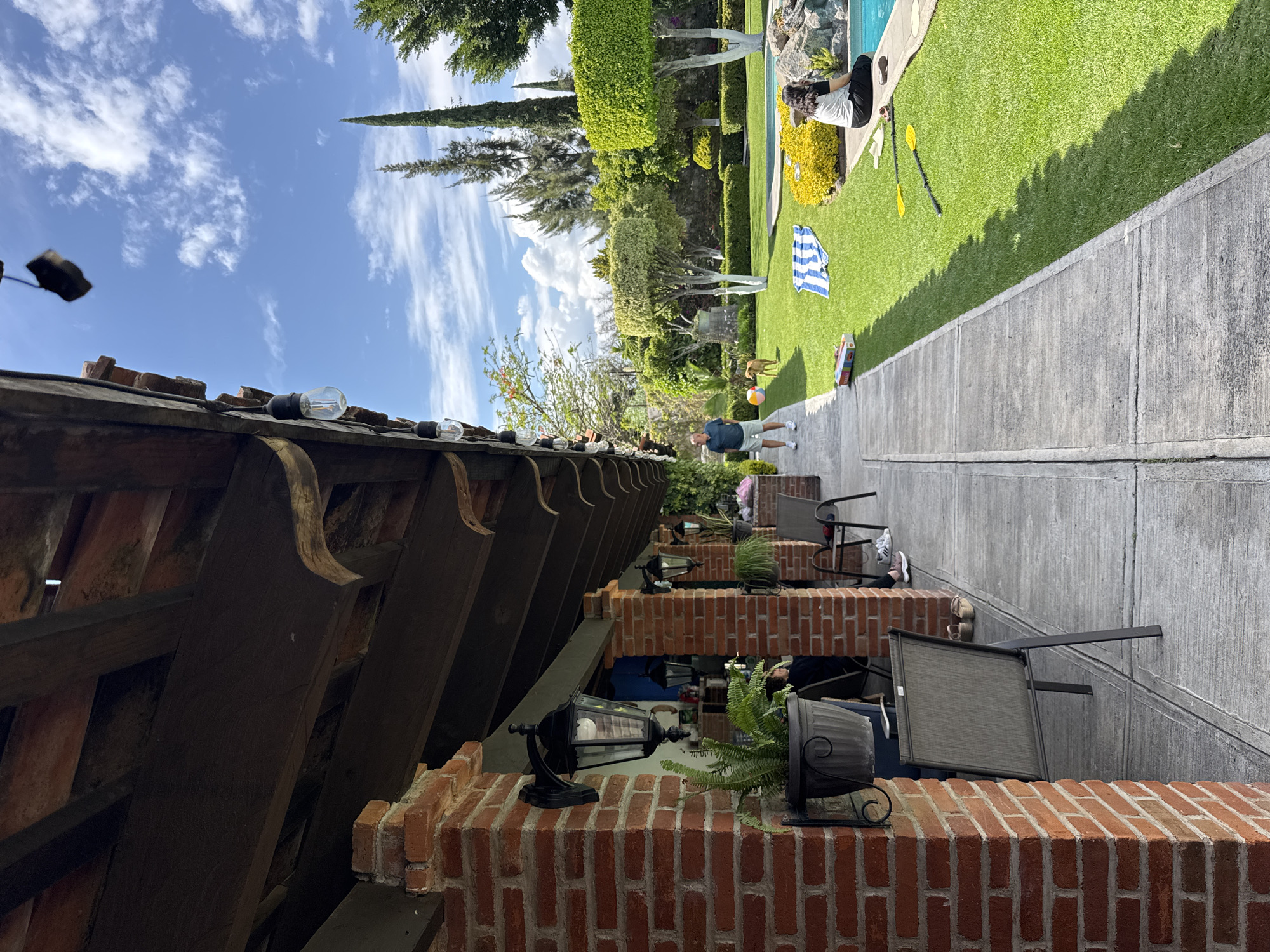The scene is an outdoor space in Puebla, Mexico, featuring a brick structure with hanging lanterns and potted ferns in the foreground. Behind it, a concrete walkway leads to a green lawn area. On the lawn, a person is seated by a swimming pool while another person is playing with a dog, throwing a ball to it. A blue and white striped towel lies on the grass, and there is a children's toy or game box near the edge of the lawn. The sky is partly cloudy.