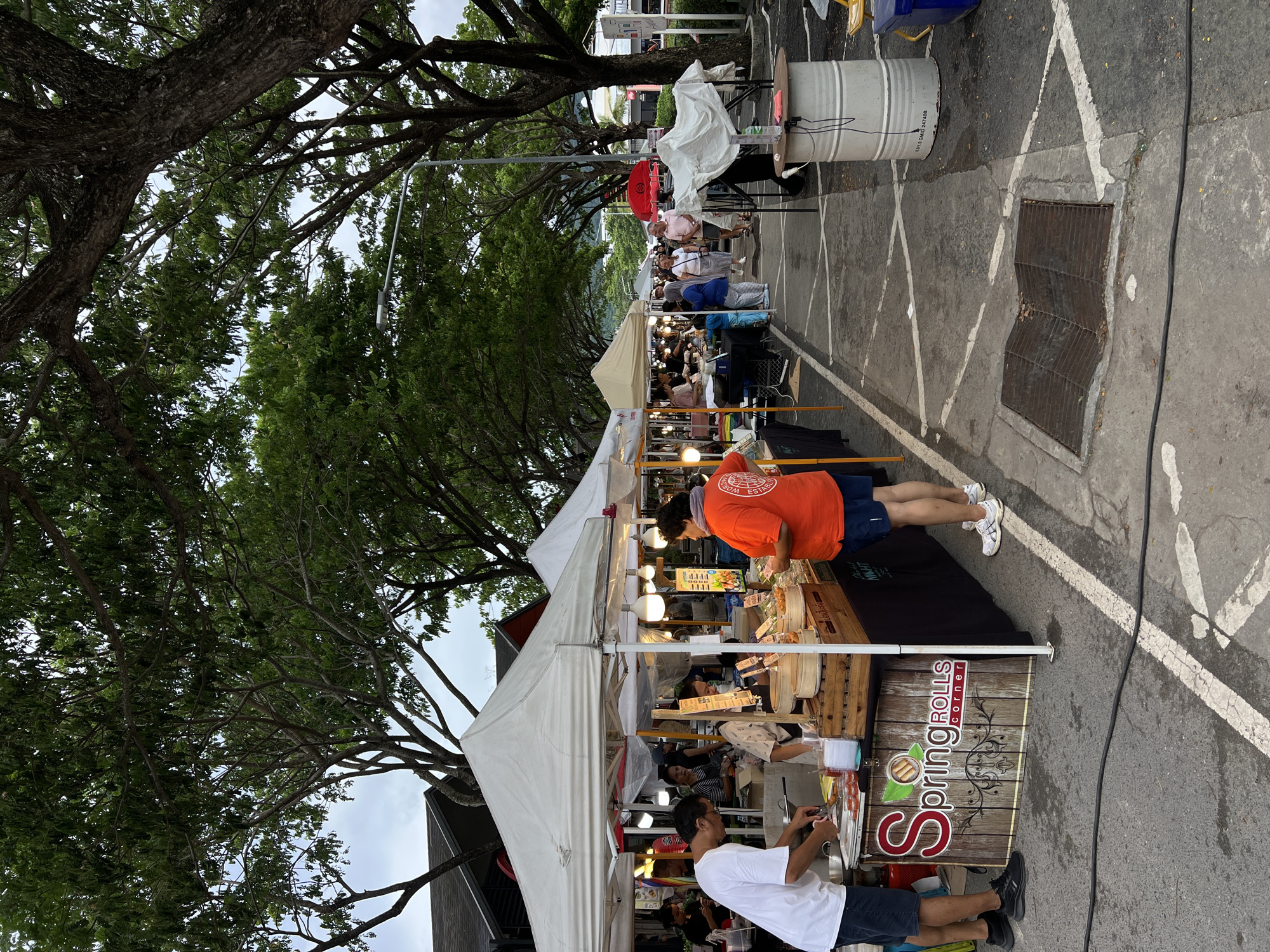 A street market is taking place under a canopy of mature trees. A variety of tents and stalls line a paved walkway. In the foreground, a vendor in a white t-shirt is preparing food at a stall labeled "Spring Rolls Corner". Another person in an orange t-shirt is standing at the same stall. Several other individuals are browsing at nearby stalls further down the walkway. The scene appears to be an outdoor market with a variety of food and goods being sold.