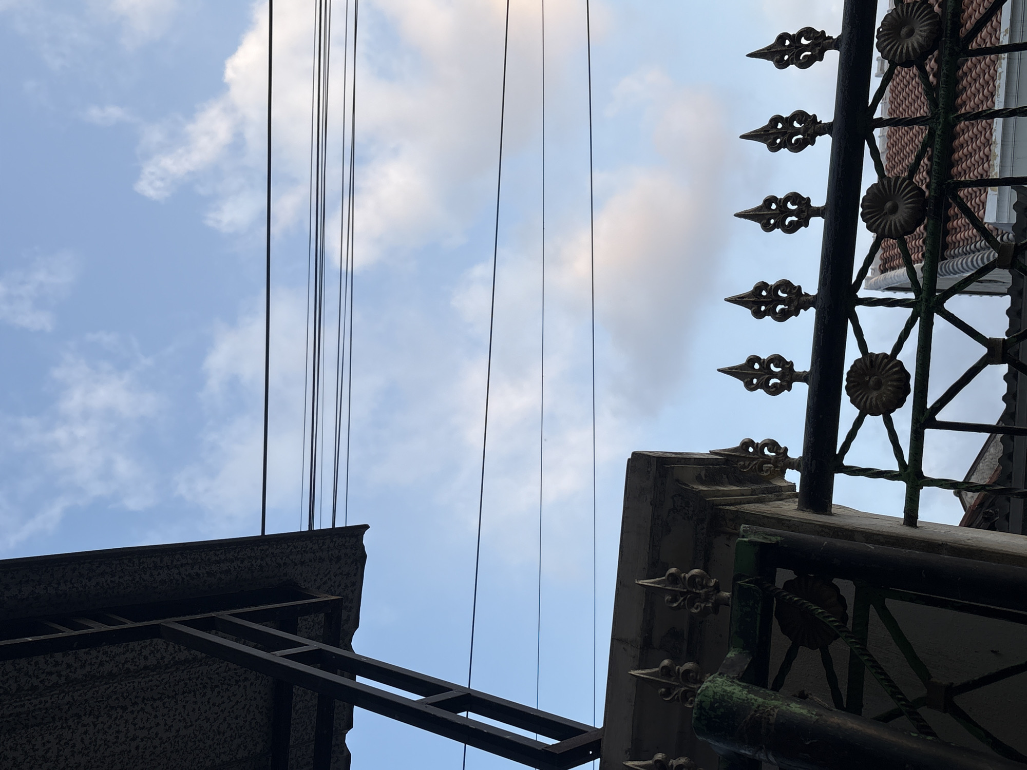 The image shows a view looking upwards towards a blue sky with scattered white clouds. In the foreground, a dark, ornate metal fence is visible, topped with decorative finials. To the left, a portion of a dark, sloped roof structure with visible supports is present. Several black electrical wires are stretched horizontally across the frame against the sky. The fence appears to be old and shows signs of wear, with some green patina visible on the metal. In the background, a tiled roof of a building can be seen through the fence. The scene depicts an outdoor environment, likely a residential area in Palimanan, Indonesia, with elements of urban infrastructure and architectural details. There are no people visible in the image.