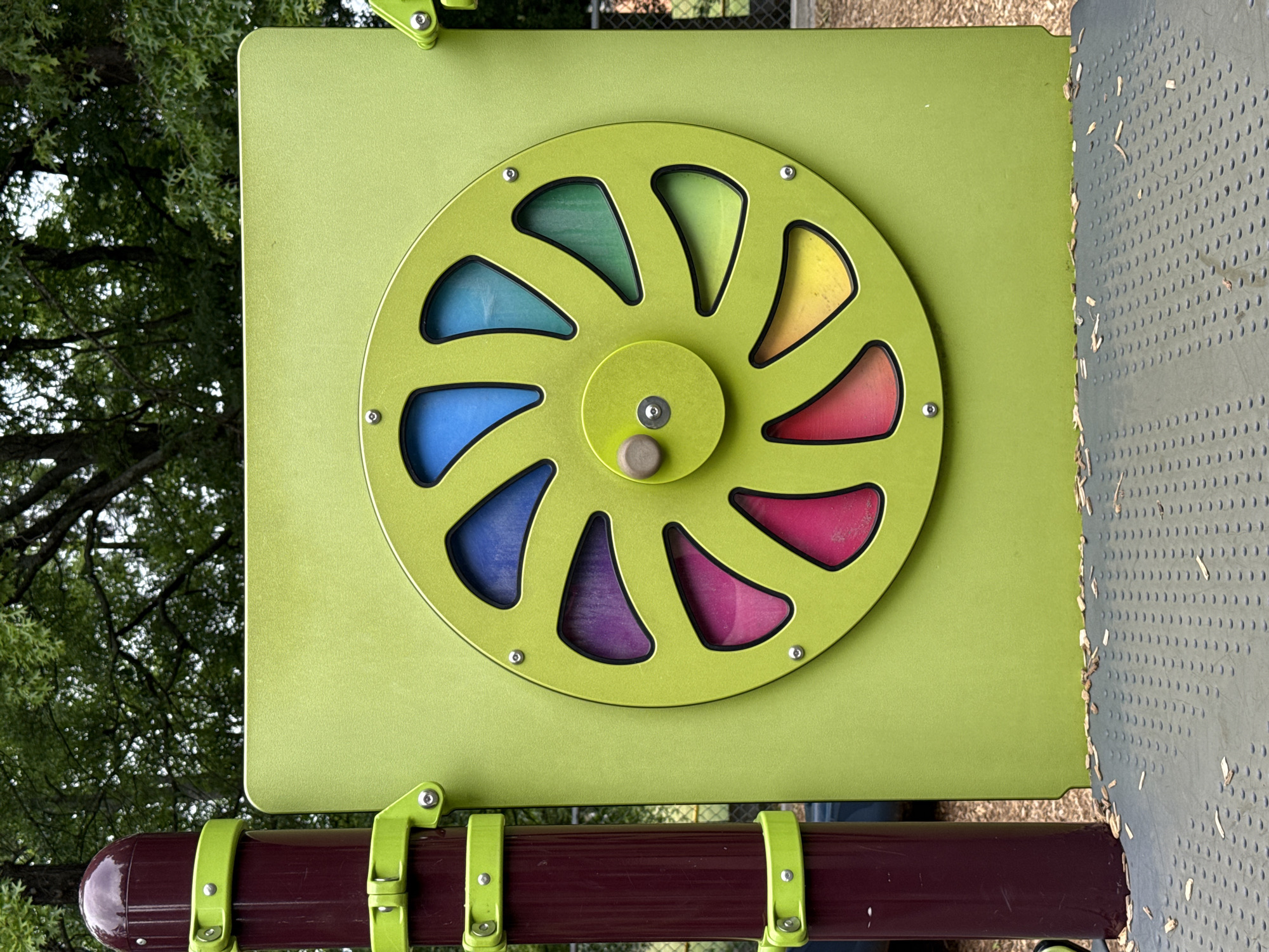 The image shows a colorful spinning wheel activity on a playground in Mountain Park, Georgia, United States of America. The wheel is circular with multiple triangular openings filled with different colors, mounted on a green square panel. The panel is attached to a red railing with green supports. The wheel is a green wheel with different colored triangular sections, including blue, purple, red, orange, and yellow. The play equipment is in an outdoor setting near a tree and a grey metal platform with many perforations.