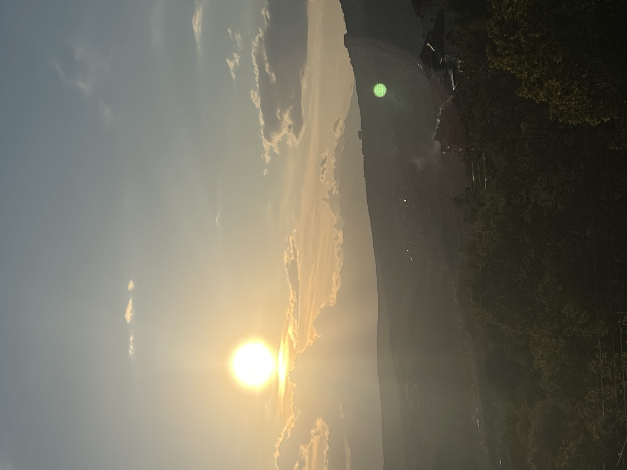 The image shows a landscape during sunset or sunrise. The sun is positioned in the lower left quadrant, emitting a bright glow. Clouds are partially illuminated by the sun's light, forming an arc towards the upper center of the image. A mountain ridge occupies the right side, with some indistinct buildings and possibly dense vegetation towards its base. The overall environment appears hazy or misty.
Without more contextual information, such as clear landmarks or features unique to Kribul, Bulgaria, it is difficult to verify the location within that city.