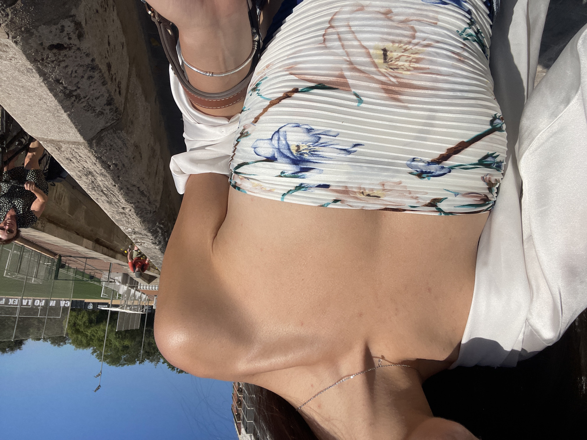 Here is a detailed description of the image:
A medium-shot captures a woman from the chest up, possibly while she is seated. She is facing the right, revealing her right shoulder and upper chest. She is adorned with a delicate necklace and wears a floral-patterned top with horizontal stripes. A white blouse drapes over her right shoulder. Her accessories include bracelets on her left wrist and a brown bag strap.
The background reveals elements of València, Spain. A stone ledge is in the upper-left corner. In the distance, a blurred figure in red is visible near a sports field. Green trees and a blue sky are present.