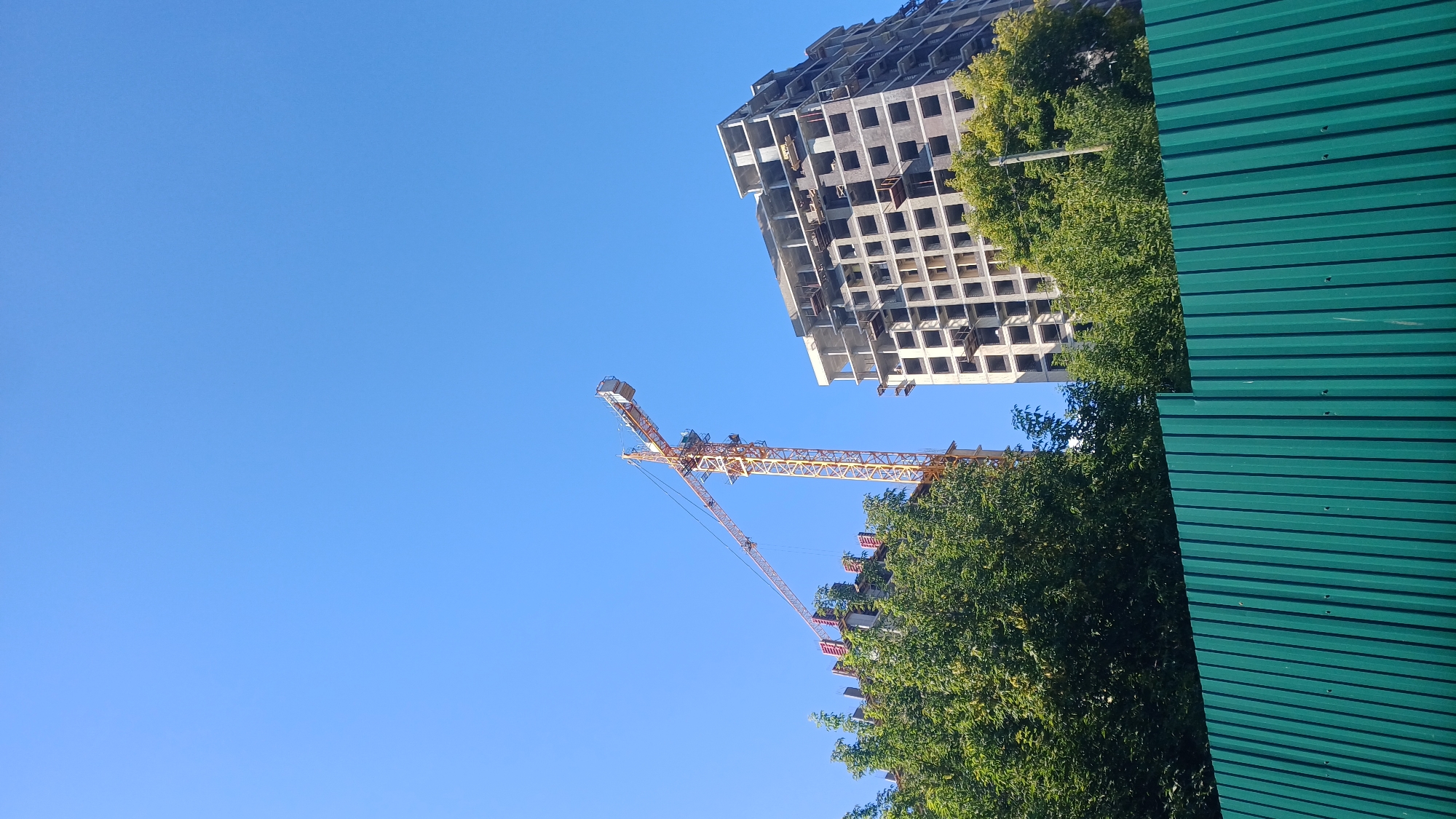 The image depicts a construction site under a clear blue sky. A tall yellow tower crane is positioned on the left, extending its boom over two unfinished multi-story buildings. The building to the right is a concrete structure with visible floor slabs and openings for windows, indicating it is in the early stages of construction. To the left of this building, another partially constructed building is visible, with what appears to be formwork and rebar. Lush green trees are situated in the foreground and between the buildings, partially obscuring the lower sections of the structures. A green corrugated metal fence runs across the bottom of the image, demarcating the construction area. The scene suggests ongoing construction activity in an urban or suburban environment. The specific location within Izhevsk, Russia, cannot be determined from the visual information alone. There are no people visible in the image.