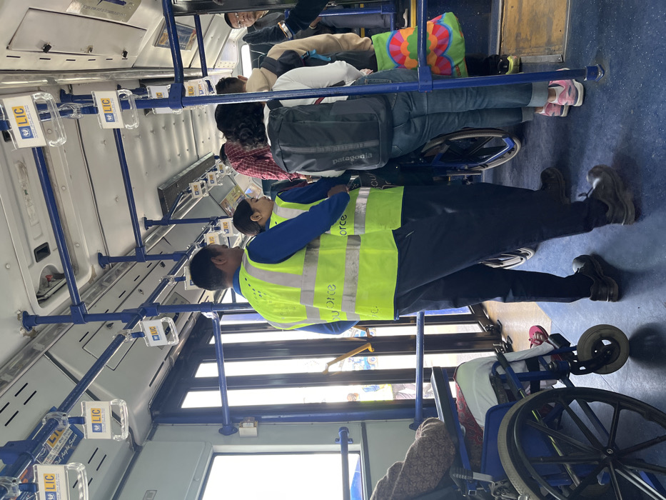 The image captures the interior of a bus, oriented sideways, with several passengers and staff members present during what appears to be daytime travel. The lighting from the windows indicates a bright outdoor environment.
In the foreground, two individuals wearing bright neon yellow high-visibility vests with reflective stripes are visible. One, facing away from the camera, is partially obscured, while the other, with shorter dark hair, is facing towards the front of the bus. The vests have text printed on them, which appears to resemble "force" or "quorce". Both individuals wear dark blue long-sleeved shirts and dark trousers.
Further back, other passengers are seated or standing. One person is comfortably seated in a blue wheelchair, which is positioned facing the front. Another person, sitting near the wheelchair, has a colorful, patterned cushion or bag. A passenger with a dark "Patagonia" branded backpack is also visible, standing or sitting. Blue handrails and support poles are prominent throughout the bus, along with a blue-colored floor.
Numerous clear plastic holders displaying the "LIC" logo (Life Insurance Corporation of India) are attached to the handrails, suggesting either advertising or sponsorship. One of these holders also has partially visible text that reads "Dal Aaple Santh," likely part of a slogan. The overall scene suggests public transport or a guided tour setting, likely in India given the "LIC" branding and the provided context of Guwahati.