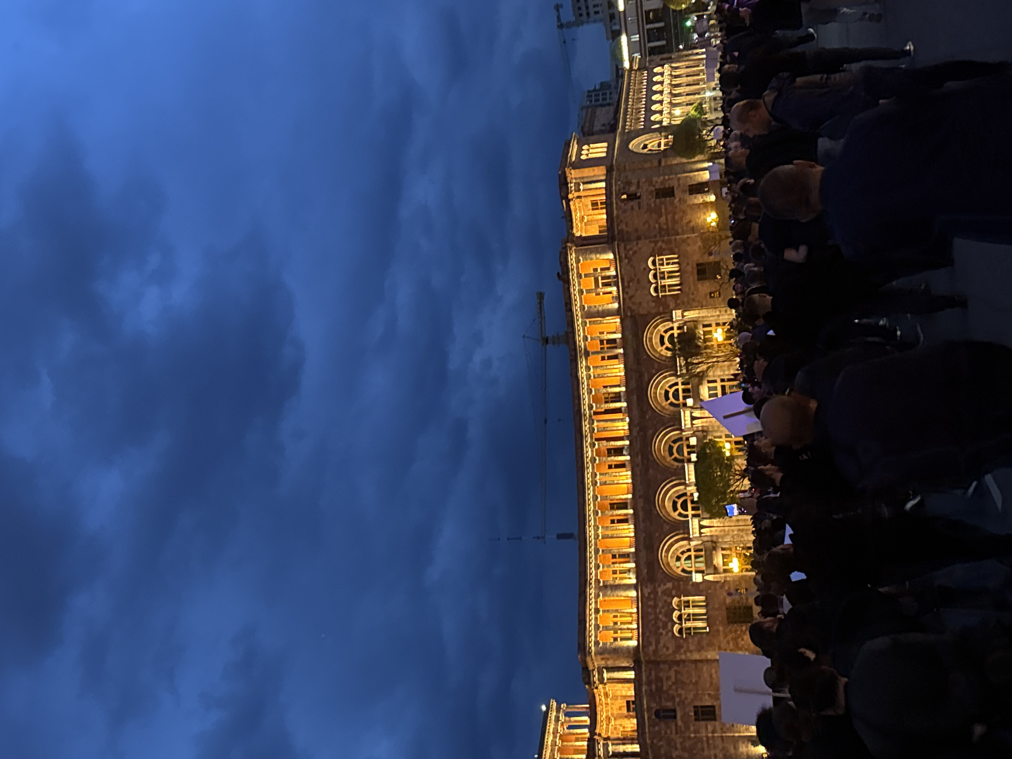 The image shows a crowd of people gathered in front of a large, illuminated building in Yerevan, Armenia. The building is multi-storied and constructed of stone with rows of arched windows. A construction crane is visible in the background. The sky is a dark blue with clouds. The crowd appears dense and mostly composed of people wearing dark clothing, some of whom are holding signs.