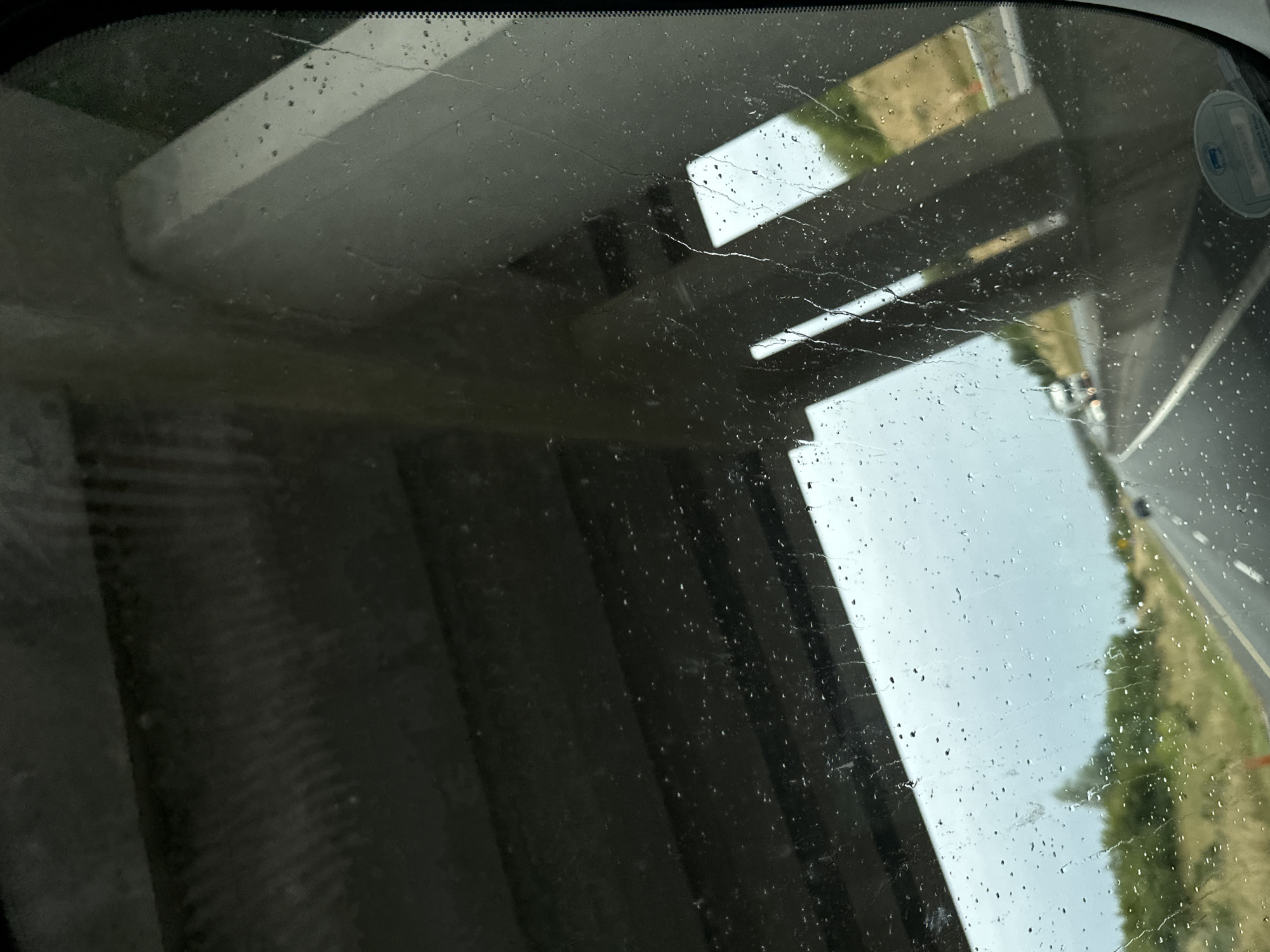 The image was taken from inside a vehicle, looking out through a rain-streaked windshield. The view is of a highway with cars in the distance. The immediate foreground is dominated by the underside of a concrete overpass, with water droplets obscuring the view. To the right, a portion of the vehicle's interior is reflected in the windshield. The overall lighting suggests an overcast or rainy day. The location is identified as Lackabeg, Ireland. There are no people clearly visible in the image.