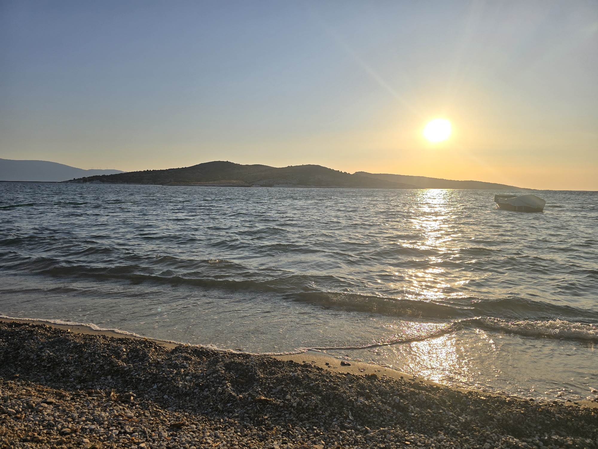 The image depicts a serene coastal scene at sunset in Foça, Turkey. The sun, a bright orb on the horizon, casts a warm, golden glow across the tranquil sea. Gentle waves lap at a pebbly shore, creating a soft white foam. In the distance, a chain of low, undulating hills forms the backdrop, their silhouettes softened by the fading light. A small, white boat, partially covered, floats placidly on the water. The sky above is a clear gradient of pale blue to soft orange, with the sun's rays creating a shimmering path on the water's surface. The foreground is dominated by the textured expanse of small, smooth pebbles that make up the beach. There are no people visible in the image.