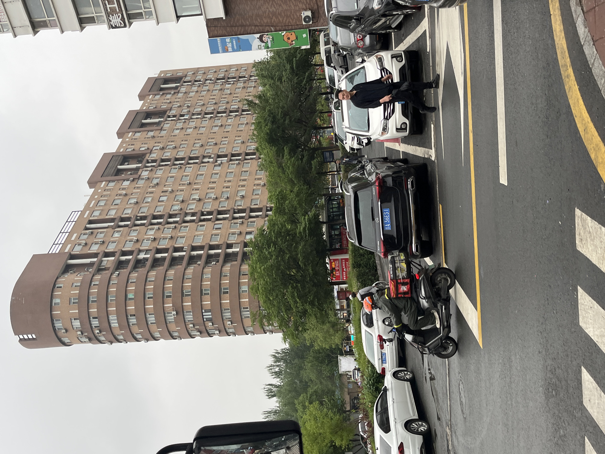 The image depicts a street scene in Changchun City, China. A tall, light-brown building with numerous windows and air conditioning units dominates the left side of the frame. The sky is overcast. A row of parked cars, mainly white and black sedans, lines the street. One black sedan displays the license plate "吉A 36ES1". A man in a black shirt and pants is standing next to a white car. Further down the street, a person on a scooter with a red package is visible. The road markings include white and yellow stripes. The license plate on the white car behind the scooter is "吉A 79MC3".