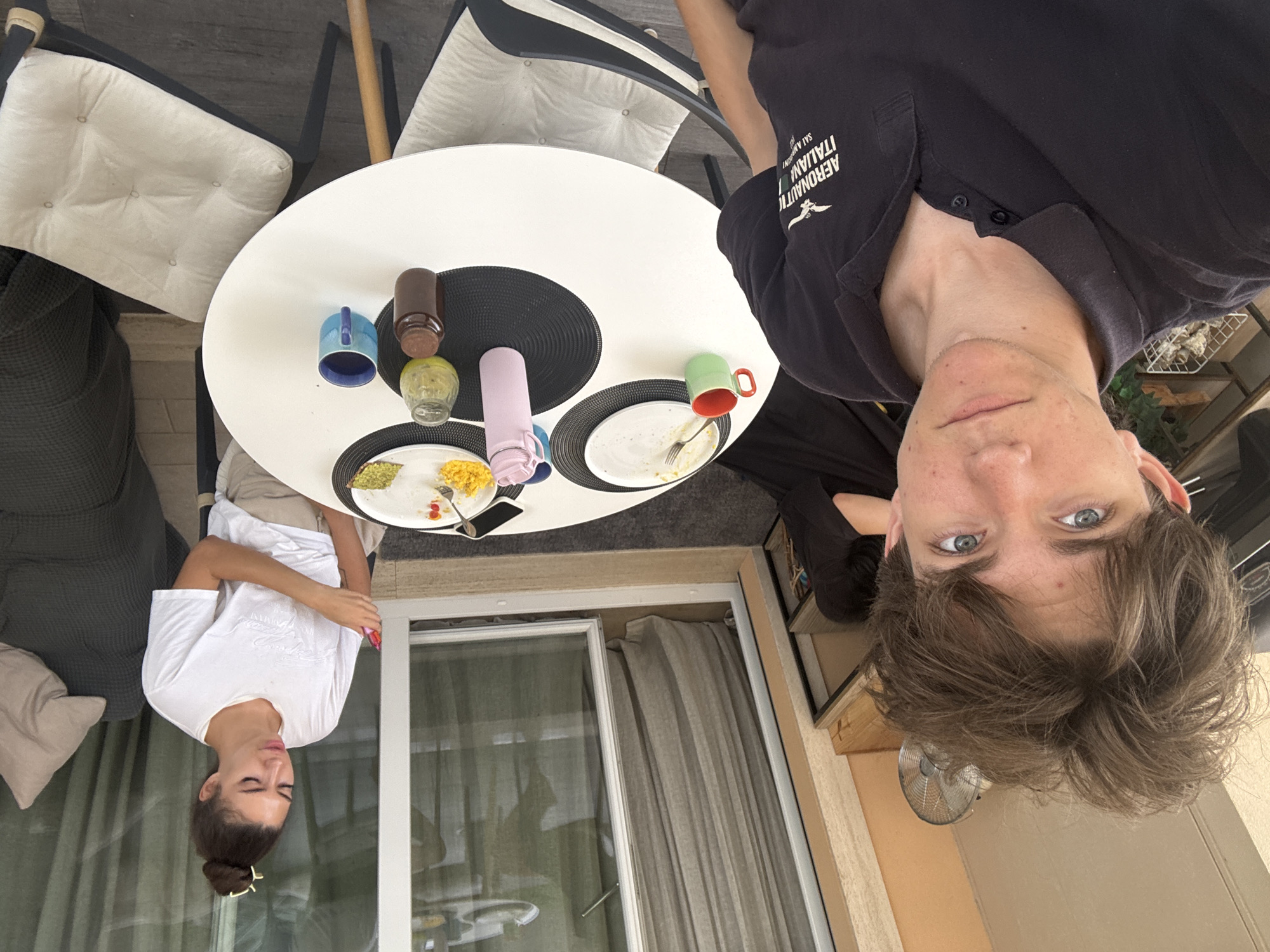 This is a media file depicting a meal with two people. One subject is a young woman wearing a white t-shirt, with her hair tied up in a bun. She is sitting at a round, white table and appears to be looking down at her plate. The other subject is a young man wearing a dark blue polo shirt with text on it. His head is tilted back, and he is looking directly at the camera. The table is set for a meal with plates of food, cutlery, and several mugs and bottles. The scene appears to be an outdoor dining area, possibly a patio or balcony, with a glass door and curtains visible in the background. The lighting suggests it is daytime.
The text visible on the man's shirt reads "AERONAUTICA MILITARE ITALIANA". The overall mood appears casual and relaxed, suggesting a meal shared between friends or family. The location context provided places the scene in Cupra Marittima, Italy.