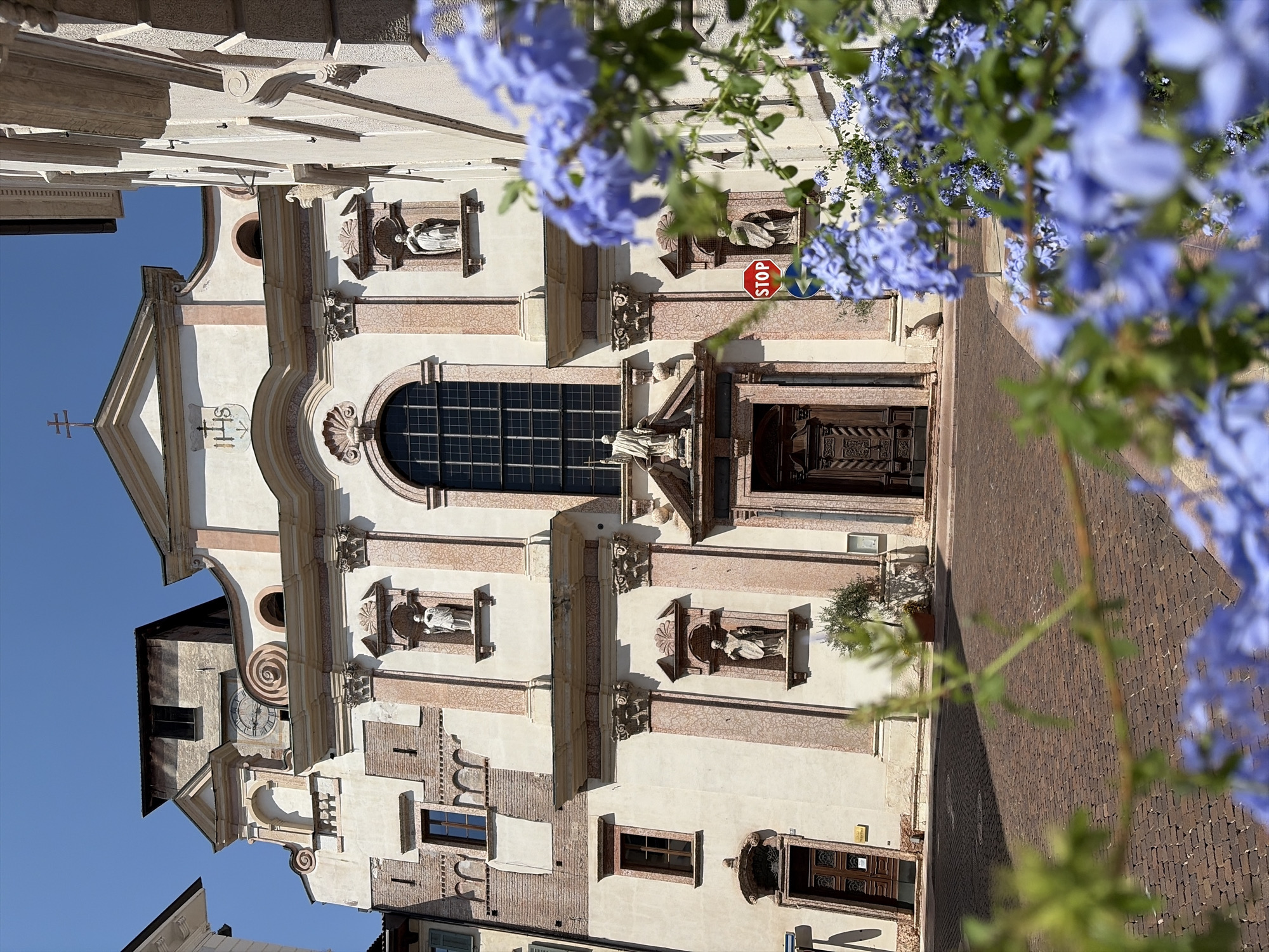 In Trento, Italy, the facade of a church is captured on a sunny day, with a clear blue sky overhead. The building features classical architectural elements such as columns, pediments, and statues in niches. A large arched window dominates the central section of the facade, above a grand wooden door. A clock tower is visible behind and to the left of the main structure. In the foreground, out-of-focus blue flowers and green foliage obscure part of the view, creating a soft, blurred effect. The street in front of the church is paved with red brick. A red "STOP" sign is affixed to a pole on the right side of the scene.