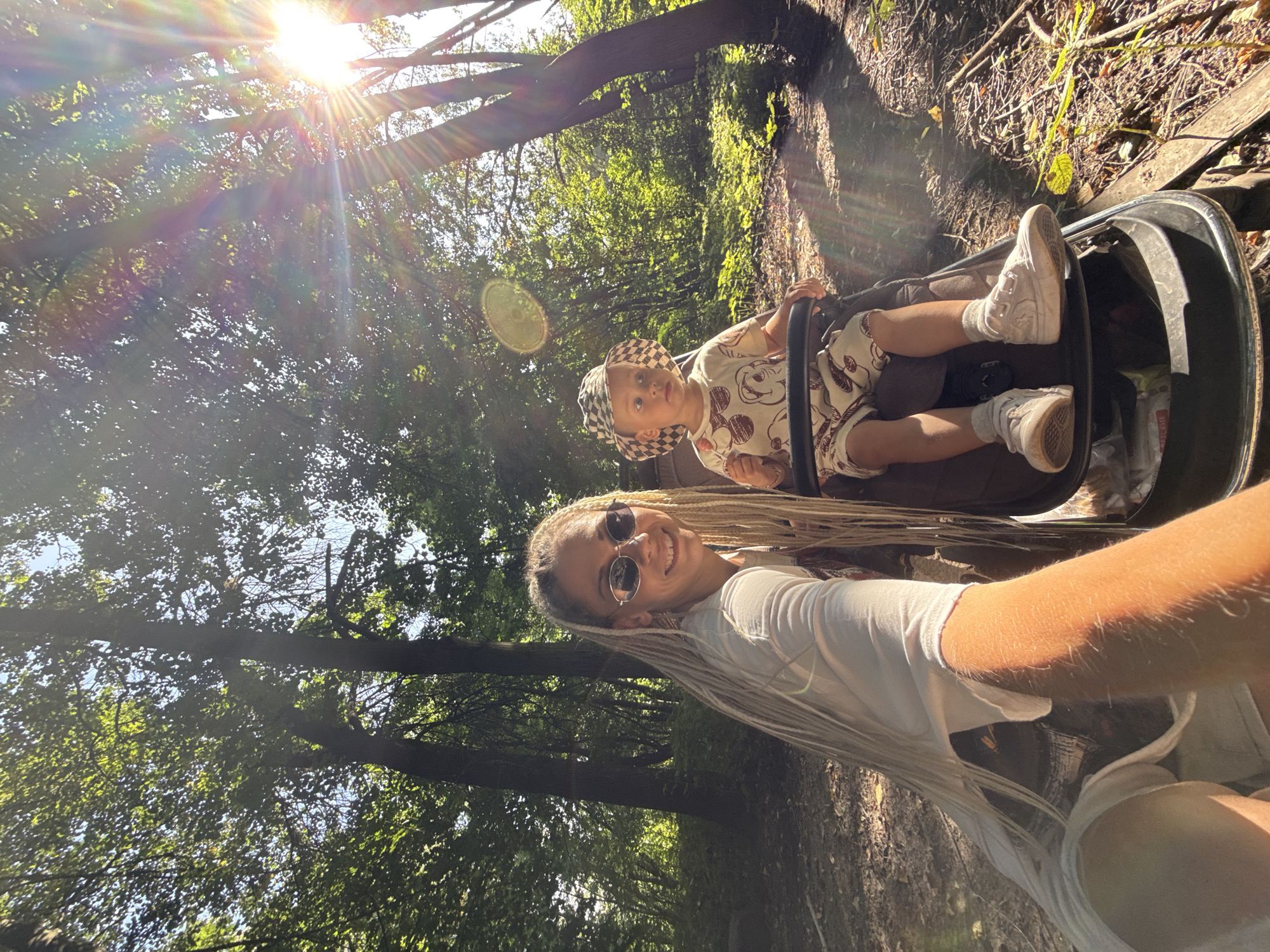 A woman with blonde braids and sunglasses is in the foreground of a selfie, smiling at the camera. She is wearing a white t-shirt and white shorts. Next to her, in a stroller, is a young child wearing a checkered hat and a light-colored outfit with a pattern of round shapes. The child is holding a lollipop. They are in a wooded area with trees and dappled sunlight. The location is the city of Rusanovo, Russia.