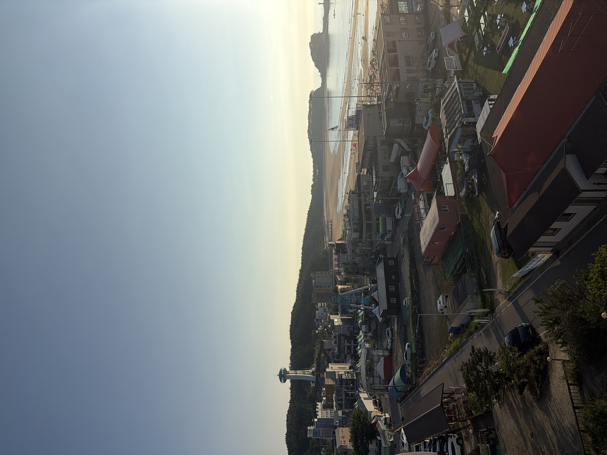 The image captures a coastal view of Mohang-ri, South Korea, bathed in the soft light of either sunrise or sunset. The scene is dominated by the vast expanse of the sky, transitioning from a clear, pale blue at the top to a warmer, yellowish hue near the horizon. Below the sky, a town sprawls towards a sandy beach and the sea.
In the foreground, a road winds through the town, lined with various structures. These include low-rise buildings, some with red roofs, and what appear to be covered market stalls or outdoor seating areas. Several cars are parked along the road and in lots. Trees and vegetation are interspersed throughout the urban landscape.
Further into the scene, the town gives way to a wider beach, dotted with small figures of people. The water is calm, and a few distant islands are visible on the horizon. A prominent lighthouse stands to the right of one of these islands.
Dominating the cityscape, on a hill overlooking the town, is a tall, slender tower with a circular observation deck at its top, a distinctive landmark. Buildings of varying heights and architectural styles populate the town, including some multi-story structures that suggest commercial or residential use. Tents and temporary structures are also present, hinting at a possible event or seasonal setup. The overall impression is one of a peaceful, seaside community during a quiet part of the day.