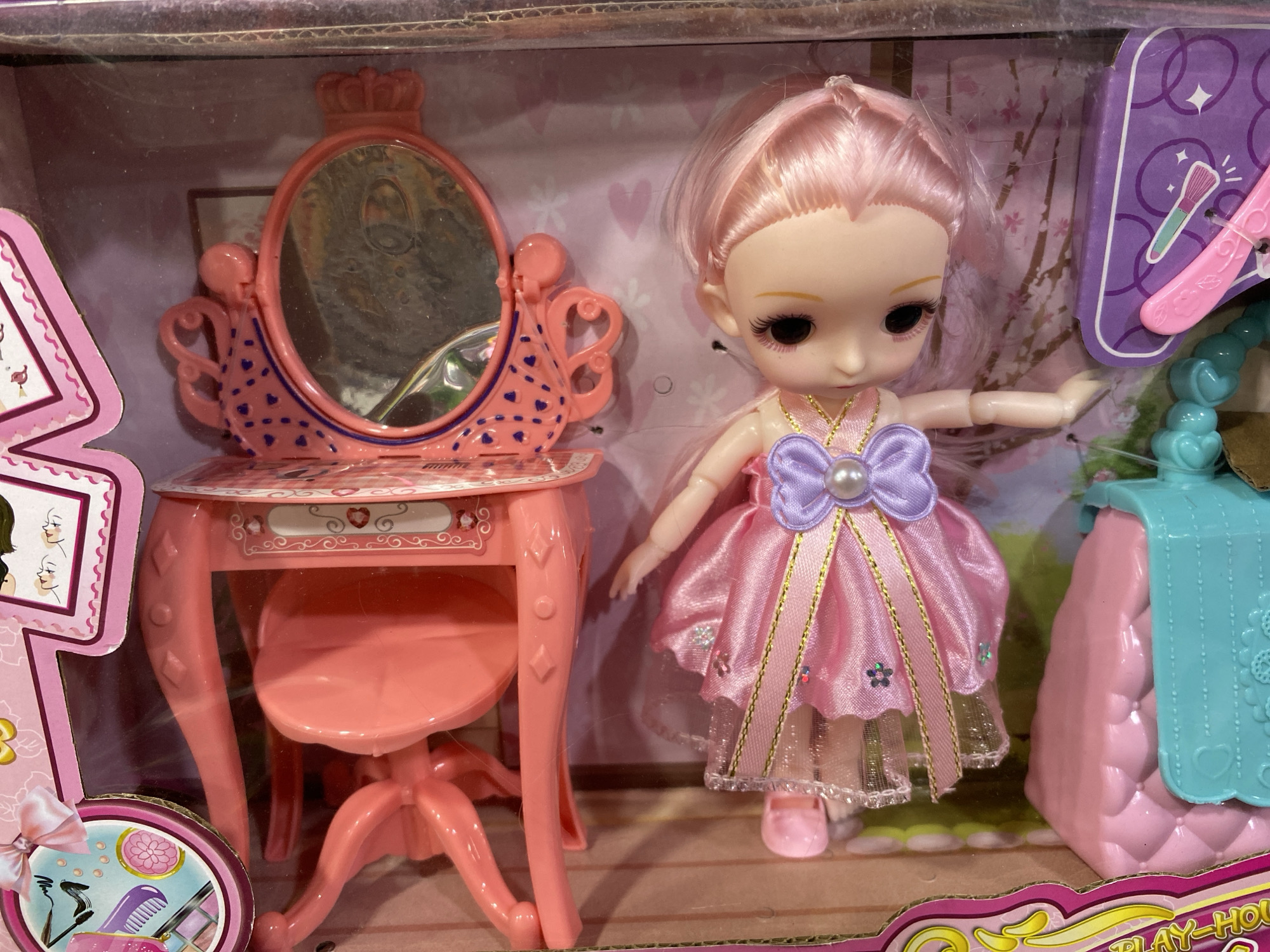 A pink doll with pink hair is standing next to a pink vanity table and stool. The doll is wearing a pink dress with a purple bow. The vanity table has a mirror with a crown on top and decorative hearts. To the right of the doll is a pink purse with a turquoise flap. The toys are in their packaging, which appears to be a cardboard box. The background of the packaging is pink and decorated with floral patterns and hearts. The scene is likely a toy store display. The location is not specified in the image, and therefore it cannot be determined to be in Banda Aceh, Indonesia.