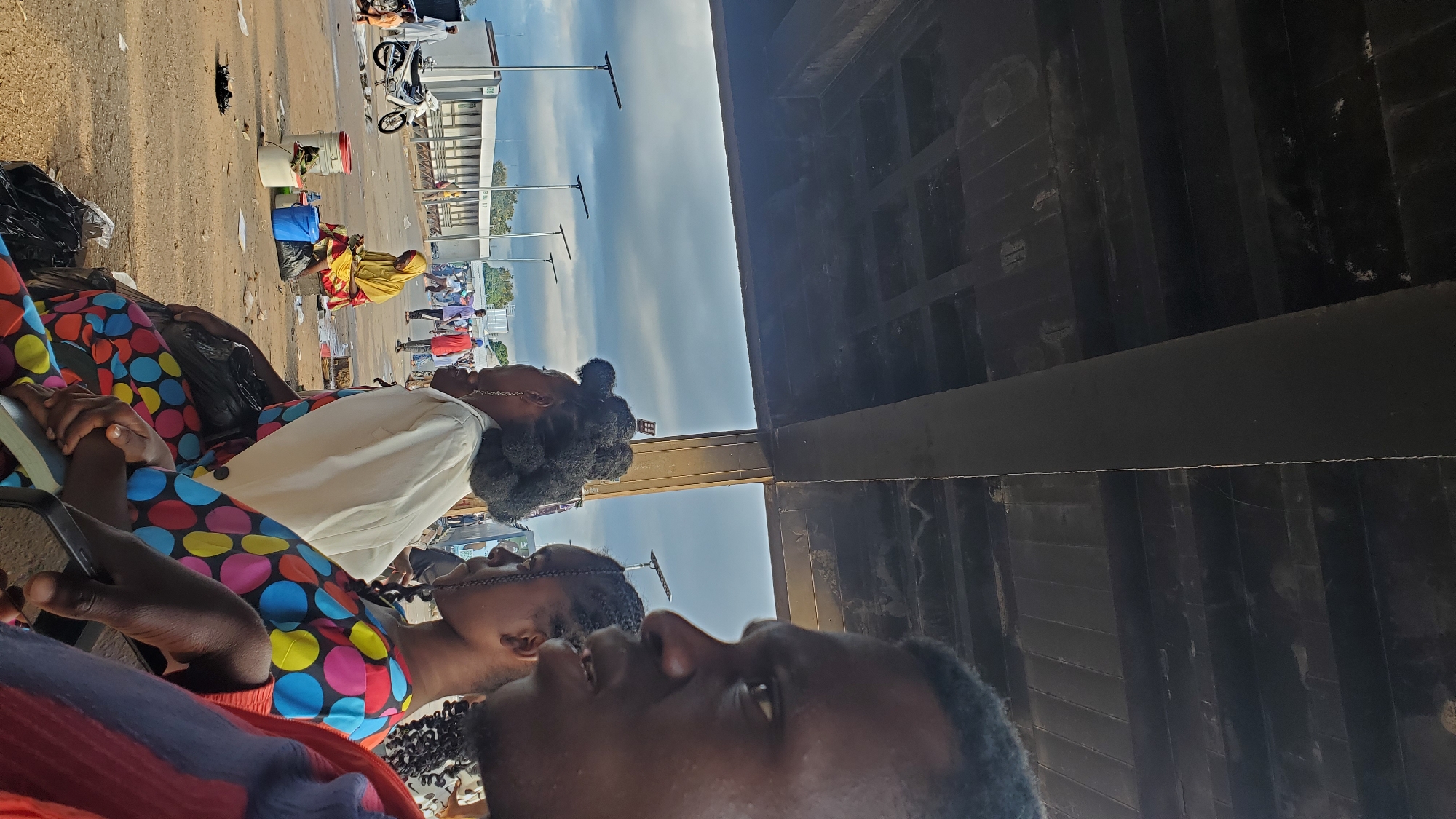 A close-up, medium shot captures a group of people sitting outdoors under a dark, textured concrete structure. In the foreground, on the right, a person with dark skin, wearing a red and blue striped garment, is partially visible from the chest up, looking to the left with their mouth slightly open. To their left, a young person with dark skin and braided hair wears a brightly colored polka-dot dress. Next to them, another person with dark skin and voluminous, dark curly hair wears a white jacket over their shoulders. This person also wears a long, dark skirt and a gold earring. In the mid-ground, a person in a bright yellow hijab and a patterned skirt is seated on the ground, looking down at something in their lap. They are surrounded by various containers, including a blue bucket and white buckets with red lids, as well as a black plastic bag. A white motorcycle is parked to the left of this person. In the background, several people are walking, and a white building with a series of openings is visible. Streetlights with dark poles are scattered throughout the scene. The ground is dusty and appears to be dirt or gravel. The sky above is cloudy and overcast.