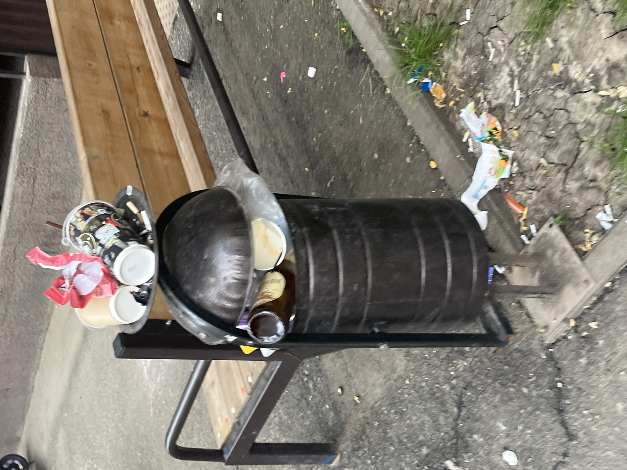This image captures an outdoor urban scene in Krasnodar, Russia, featuring a severely overflowing public trash can positioned next to a wooden bench. The dark, cylindrical metal trash can, supported by a dark metal frame and topped with a domed lid, is completely overfilled with refuse. A round, flat metal tray, functioning as an ashtray, rests on the domed lid, holding several used disposable coffee cups (some light brown, others with graphics), a crumpled red and white wrapper, and multiple cigarette butts. Within the primary opening of the trash can, a brown glass bottle, additional white paper cups, and a clear plastic bag are visible.
Behind the trash can, a wooden bench with light-colored slats for the seat and backrest, supported by dark metal armrests and legs, is present. The ground in the foreground and extending to the right consists of dark gray asphalt or concrete, exhibiting numerous cracks, particularly on the right side. This ground area is heavily strewn with various pieces of litter, including numerous cigarette butts, small white fragments, yellow particulate matter, a pink plastic item, and an opened white plastic bag containing colorful debris. Along the right edge of the image, a narrow strip of dry, cracked soil with sparse green grass borders a concrete curb. A small portion of a wheel is partially visible in the upper left quadrant. No individuals are present in the scene.