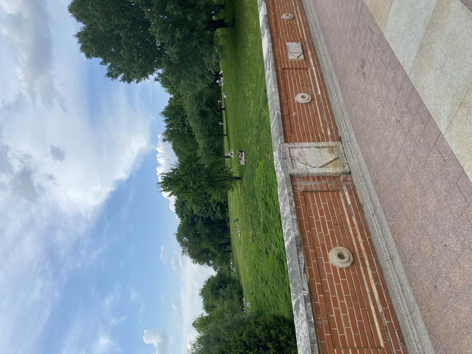 The image captures an outdoor scene in Unknown, Unknown, featuring a brick structure in the foreground. Adjacent to the brick structure is a paved area with what appears to be light-colored stone and red-brown tiles. Beyond this paved area, a large expanse of green grass stretches out, punctuated by trees. People are scattered on the grass, some seemingly resting or lounging. The sky overhead is blue with white clouds.