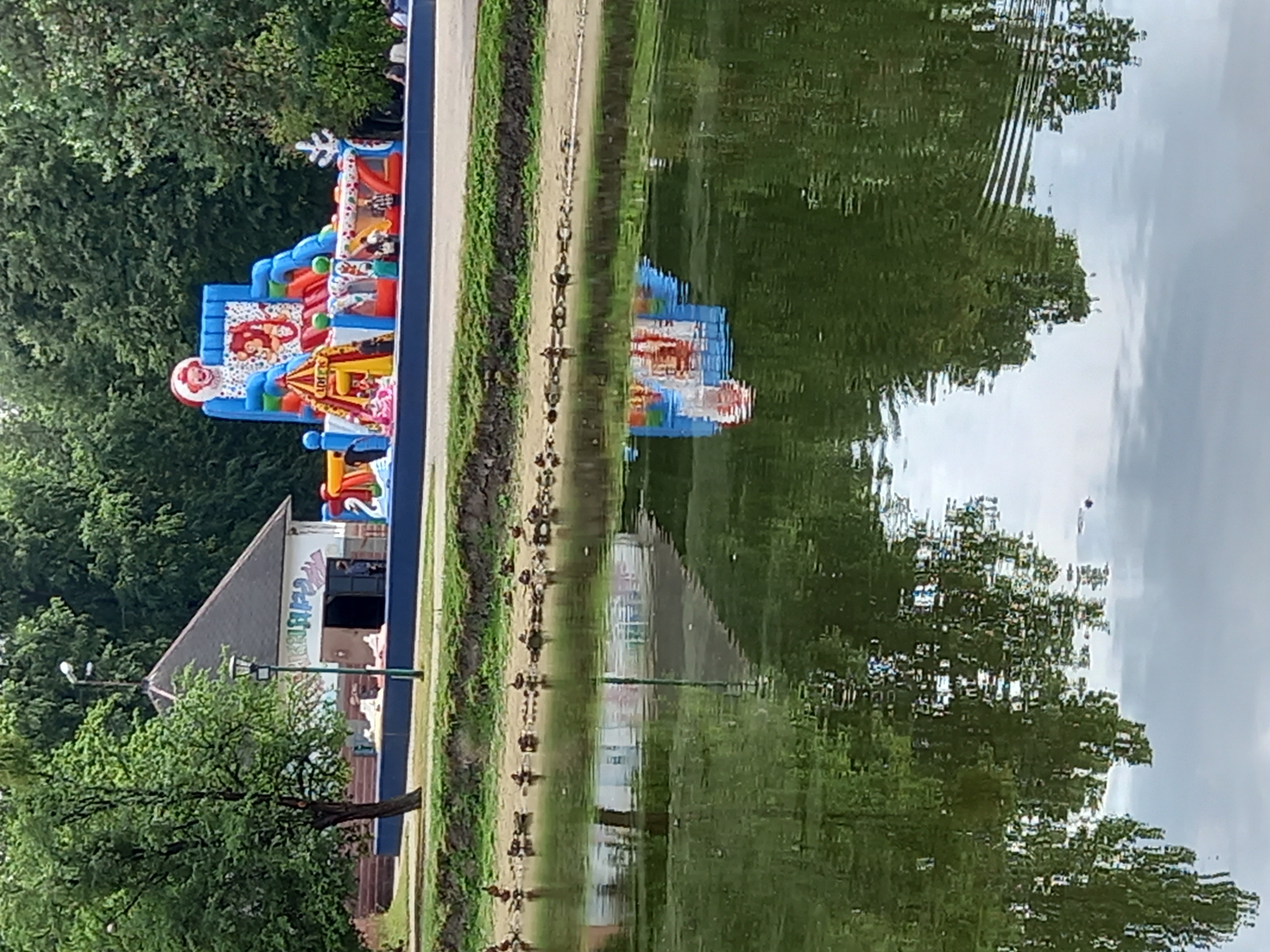 The image depicts an outdoor scene, likely a park or recreational area, as evidenced by the presence of lush green trees and a body of water reflecting the surrounding environment. The water's surface is rippled, creating distorted reflections of the trees, sky, and an inflatable playground.
The inflatable playground is a prominent object, brightly colored in red, blue, and yellow, featuring various sections and what appears to be cartoon characters. It is situated on the left side of the frame, partially obscured by trees. Adjacent to the playground is a small building with a triangular roof, possibly a concession stand or facility. Some graffiti is visible on the building's exterior. A single tree stands in front of the building, its branches spread wide.
The reflection in the water shows a distorted image of the inflatable playground, the building, and the trees. The sky appears overcast, suggesting a cloudy day. The overall impression is one of a casual, possibly family-oriented outdoor setting, perhaps during a fair or event. No specific time of day can be determined with certainty, but the lighting suggests daytime.
The provided location context is Brest, Belarus. There is no visible text in the image that can be clearly read, though some graffiti on the building might contain text.