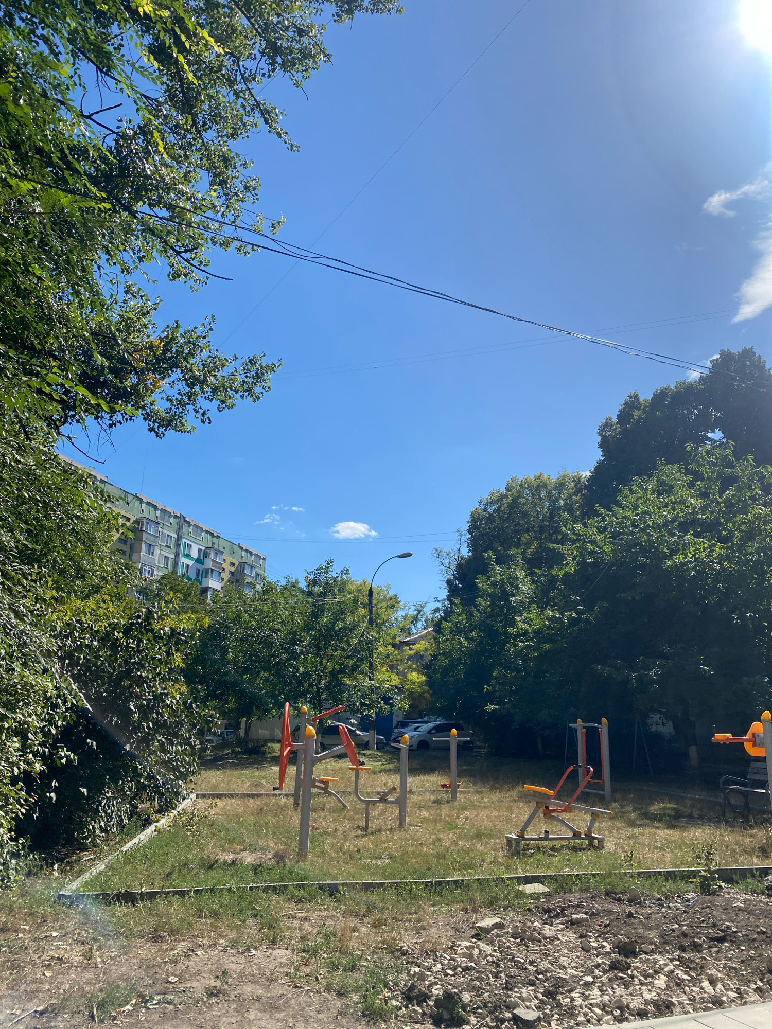 The image depicts an outdoor exercise area with several pieces of equipment in a grassy, somewhat unkempt yard. In the background, a tall, multi-story apartment building with a light green and white facade is visible, surrounded by dense green trees. The sky is a clear, bright blue with a few small white clouds. Power lines are strung across the sky. The ground in the foreground is rough, with patches of dry grass and loose stones. The lighting suggests it is a sunny day. The location is identified as Chișinău, Moldova.
