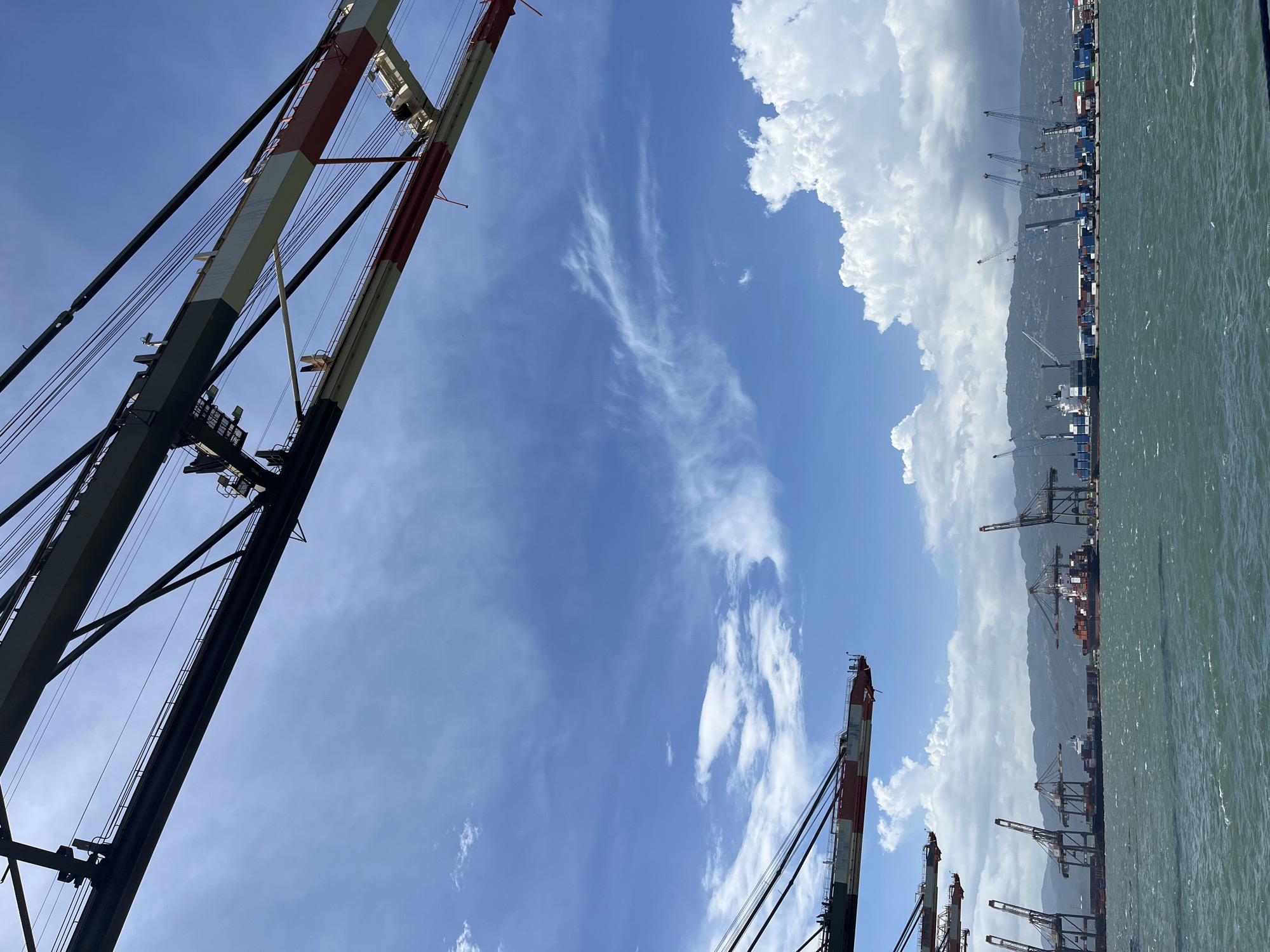 The image depicts a port in Unknown, Unknown. Cranes are visible in the foreground and background, with the sky showing a mix of blue and white clouds. Ships are docked at the pier, presumably for loading and unloading cargo. The water is relatively calm.