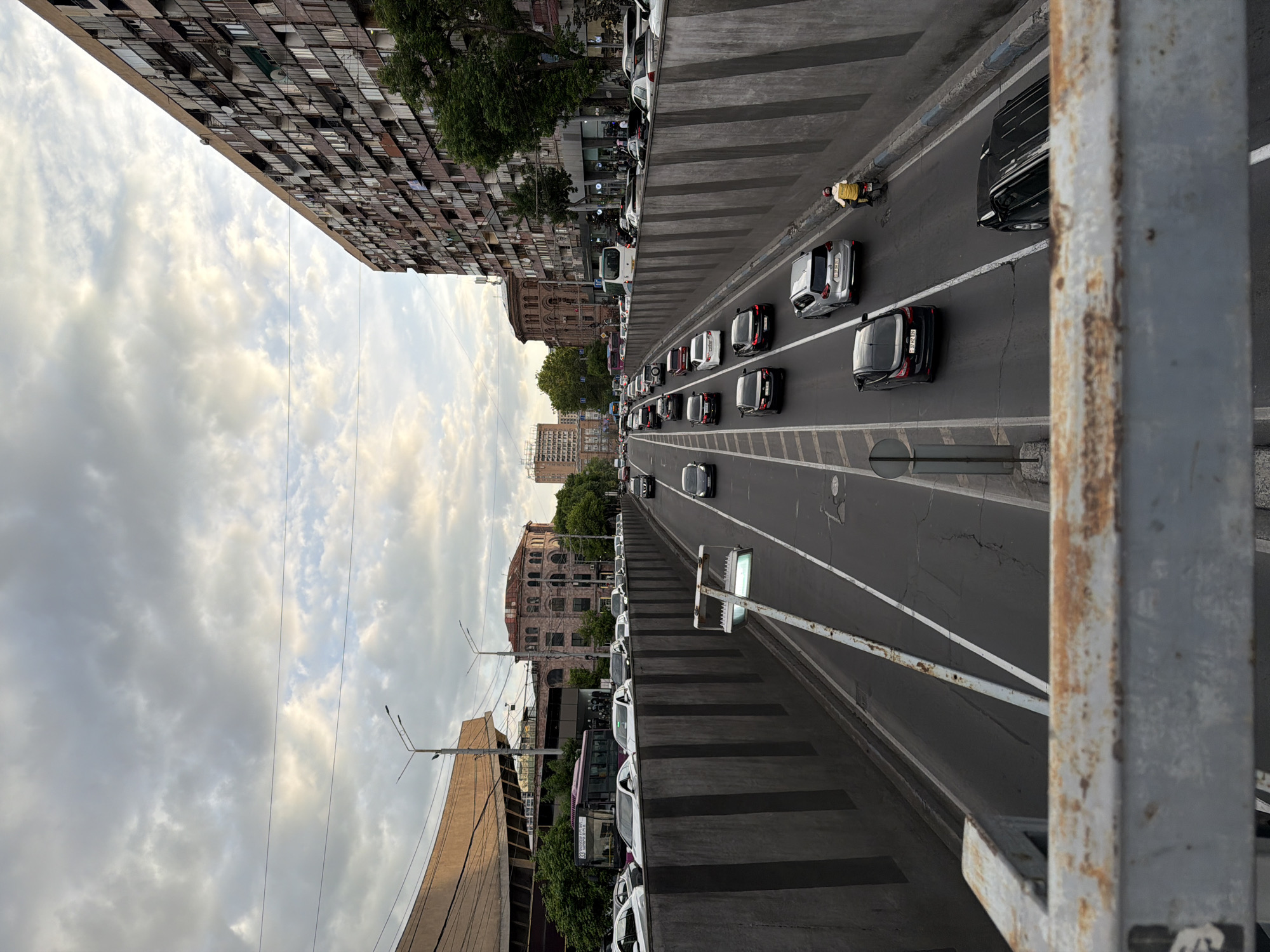 The image depicts a view of a street in Yerevan, Armenia, from an elevated position, possibly a bridge or overpass. The street has multiple lanes of traffic with cars traveling in both directions. The street is lined on both sides with buildings of various architectural styles and heights, including residential apartments, historical buildings, and modern constructions. The sky is overcast.
On the left side of the image, a tall, multi-story apartment building dominates the scene, its facade characterized by numerous windows and balconies. Adjacent to the building, a large tree adds greenery to the urban landscape.
On the right side of the street, vehicles are parked along the curb. A bus is visible among the parked cars.
The road itself shows signs of wear, with cracks and visible repairs. A cyclist is visible to the right of the road.