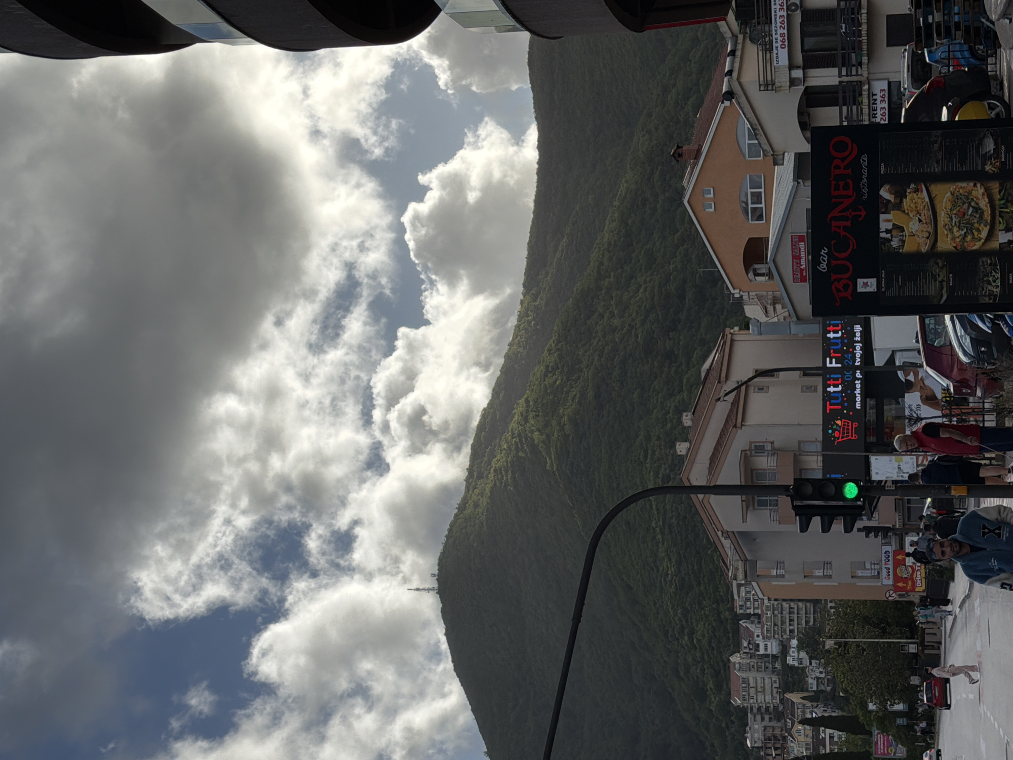The image depicts a street scene in Budva, Montenegro, with a mountain rising in the background. The sky is partly cloudy, with a mix of dark and light clouds. A street runs through the image, with buildings lining both sides. There are several commercial signs visible, including "BUCANERO," "Tutti Frutti," and "Good FOOD Drive Ta." A traffic light is green. People are walking on the sidewalk, and cars are parked along the street. Some of the pedestrians appear to be engaged in normal street activities. The overall scene conveys a typical urban environment in Budva, set against the backdrop of a mountain landscape.