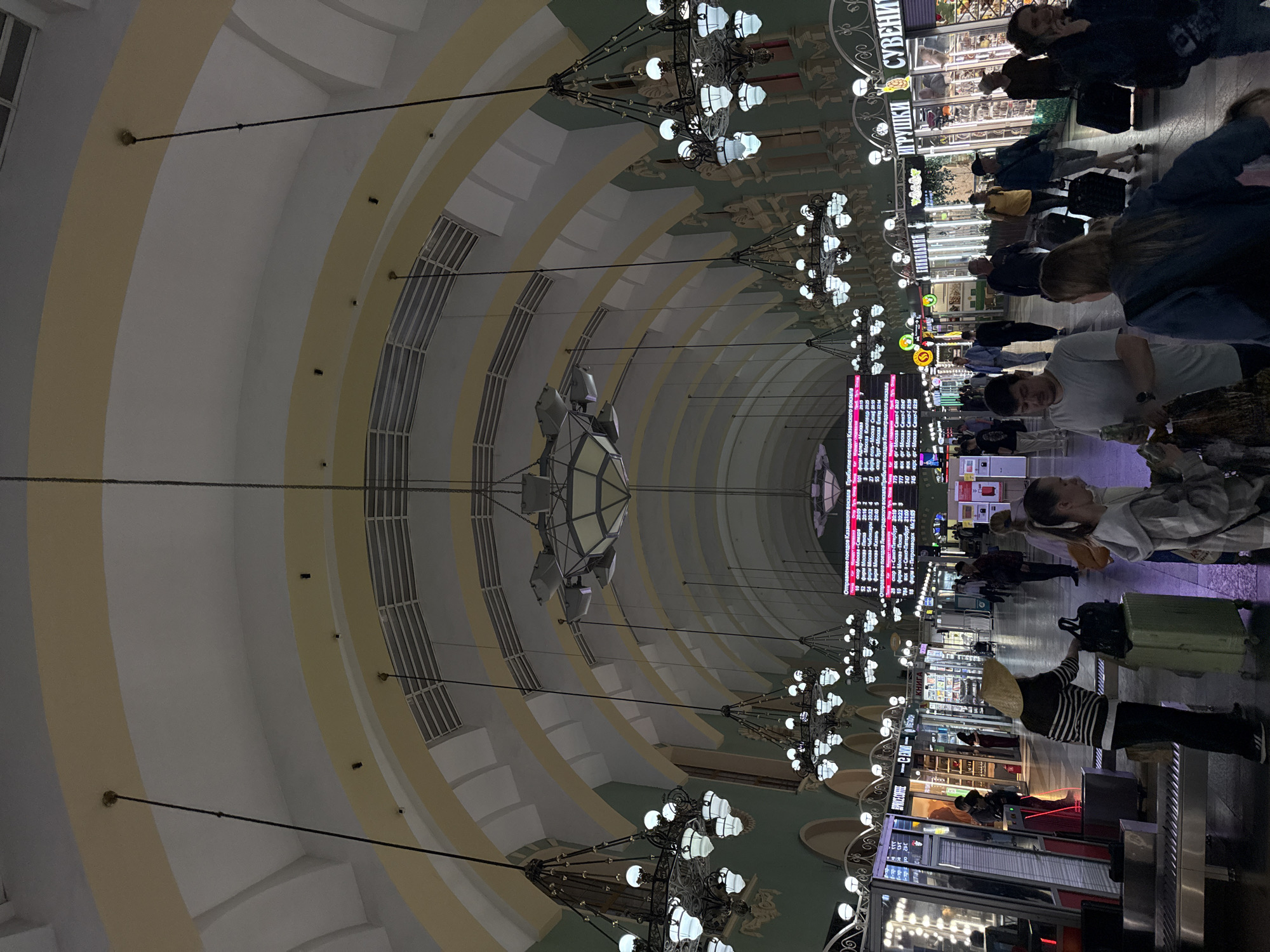 Here is a detailed description of the image:
**General Description:**
The image depicts the interior of a grand railway station concourse, likely a main hall, characterized by its high, arched ceiling, ornate chandeliers, and a bustling atmosphere. The photograph is taken from a standing level, looking lengthwise down the hall, which gives a strong sense of depth and perspective. The orientation of the image is such that the ceiling dominates the upper portion of the frame.
**Environment and Architecture:**
* The ceiling is the most striking feature, featuring a series of arches decorated with pale yellow and white paint and molding. These arches create a tunnel-like effect leading down the hall.
* Large, decorative chandeliers are suspended from the ceiling at regular intervals, providing illumination and adding to the elegance of the space.
* On either side of the hall, there are shops and services catering to travelers.
* Large display boards, likely displaying train schedules and other travel information, are visible at the far end of the concourse.
**People:**
* Many people are present in the hall, either standing or walking. Some are seen with luggage, suggesting they are in transit.
* People appear to be of diverse ages and appearances.
* Some individuals are engaged in conversations, while others seem to be focused on their destinations.
**Objects:**
* Luggage is seen in various forms, including suitcases, backpacks, and bags.
* Signage for shops and services is visible, indicating the presence of businesses within the station.
* The chandeliers themselves are notable objects, contributing to the aesthetic of the space.
* Ticketing booths are visible
**Actions and Interactions:**
* People are primarily engaged in movement, either walking through the hall or pausing to consult information.
* Some individuals are likely purchasing tickets or other travel services.
**Location:**
I am unable to confirm the image's location as Ukromne, Ukraine with certainty, because this would necessitate a more thorough examination of the image's features such as the language on signs or the architectural style.