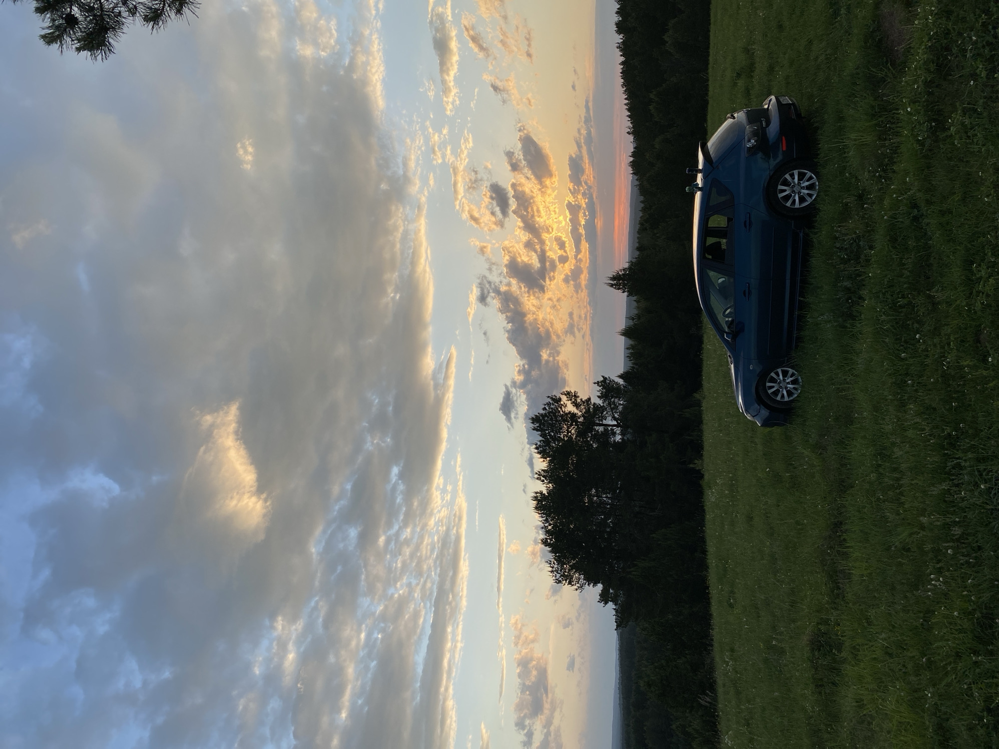 The image shows a blue car parked on a grassy field near a treeline. The sky is filled with clouds, some illuminated by the setting sun. The location is in the city of Unknown, Unknown.
