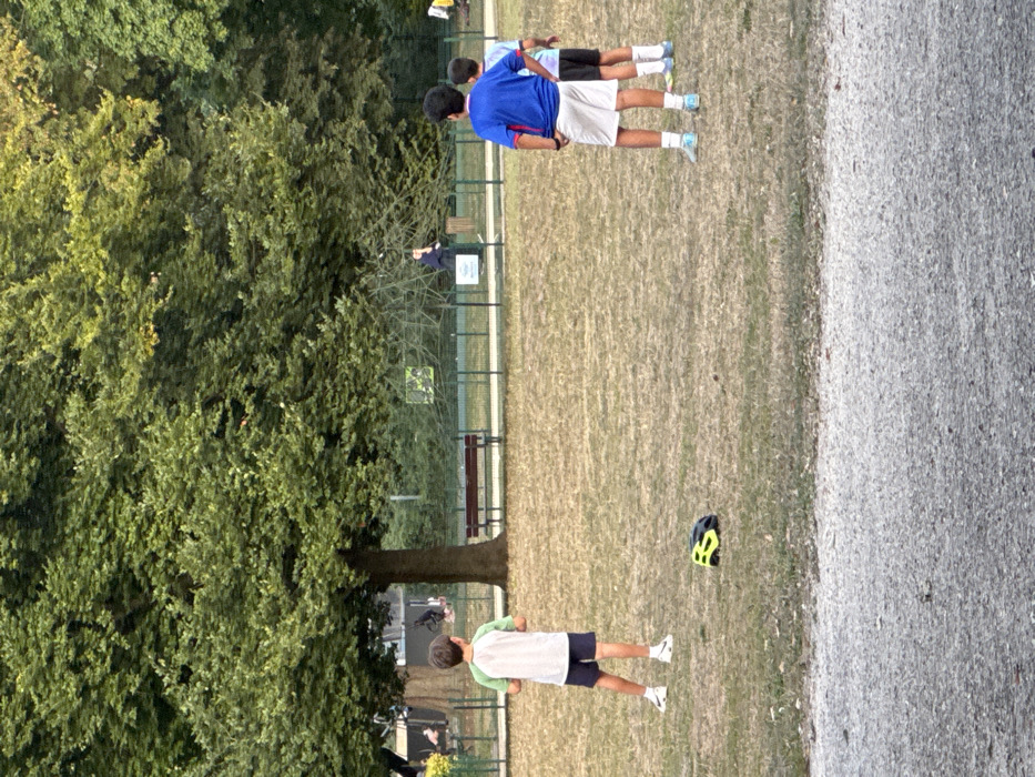 The image depicts a park scene in Conflans-Sainte-Honorine, France. The foreground consists of a gritty, light grey surface transitioning into a dry, light brown grass field. Three male children, appearing to be pre-adolescents, stand with their backs mostly towards the viewer on this field.
The child on the far left wears a white short-sleeved shirt with light green sleeves, dark shorts, and white sneakers with black Nike branding. To his immediate right, a yellow and black bicycle helmet rests on its side on the grass. The central child wears a blue short-sleeved shirt, white shorts, and light blue athletic shoes. The child on the far right is partially obscured by the central child, wearing a dark short-sleeved shirt, dark shorts, and light-colored athletic shoes. All three children are standing upright and facing away from the camera, indicating observation of an unseen element or a paused activity.
In the mid-ground, a low green metal fence separates the immediate field from a background area. Behind this fence, a dark brown slatted park bench is positioned beneath a large tree with dense green foliage. A white sign with dark, illegible text is affixed to the fence. Further in the background, a large, dark-colored climbing net structure is visible. A blurred individual wearing dark clothing is indistinctly visible near this climbing structure. The background is predominantly filled with numerous mature trees displaying green leaves, suggesting a natural park environment. The lighting is consistent with natural daylight, possibly late afternoon.