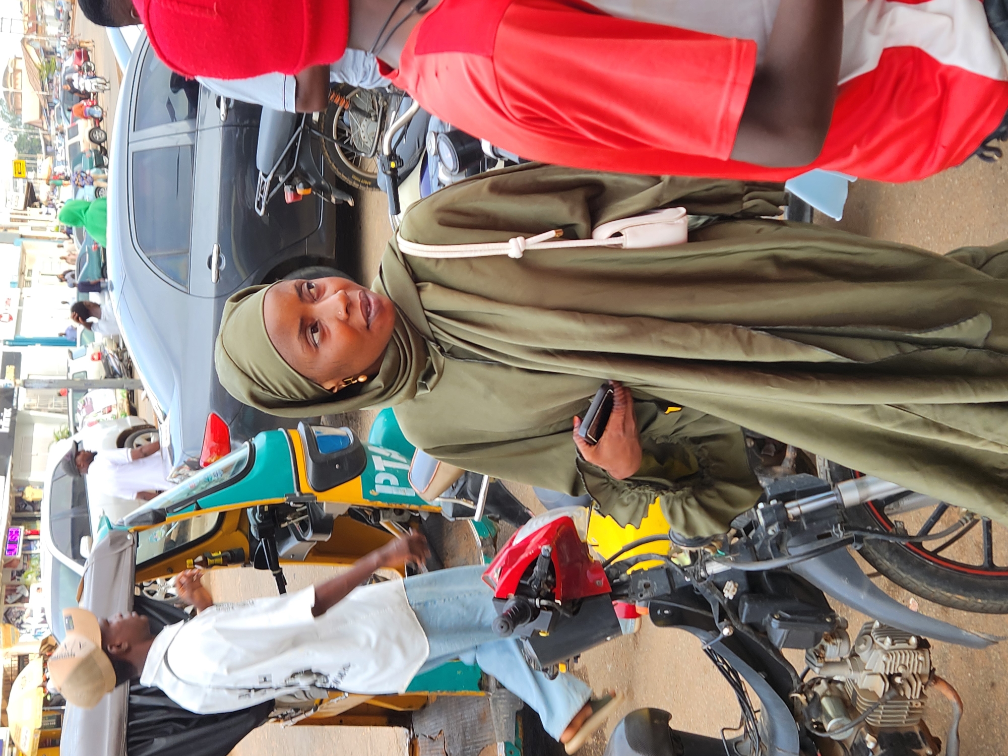 The image, rotated 90 degrees clockwise, captures a lively daytime street scene in what appears to be an urban area. The ground is unpaved, suggesting a developing locale.
In the foreground, a woman wearing a flowing olive-green khimar or hijab is prominent. She has fair skin, gold earrings, and a nose piercing. Her expression is neutral as she looks slightly upwards and to the right. She holds a black smartphone in her right hand, while a white shoulder bag with a knotted strap is visible over her left shoulder. Behind her, partially obscured, is another person wearing a red and white t-shirt, seen from the back and side.
To the left of the woman, a grey sedan car is parked, and a man in a white tunic is leaning against it in the midground. Below them, another man in a white t-shirt, light blue jeans, and a light-colored baseball cap stands, also looking upwards. He is positioned next to a distinct yellow and turquoise three-wheeled vehicle, resembling an auto-rickshaw or "keke napep," with the letters "PTA" visible on its side. Part of a motorbike's handlebars and exposed engine are visible in the lower right, suggesting a mix of transportation.
The background is busy with other vehicles, including cars and motorbikes, and blurred figures of people, indicating a bustling environment. The lighting is diffused, suggesting either an overcast day or a bright, non-direct sun. No specific weather conditions like rain are apparent. The overall impression is that of a typical, active street in a city or town, possibly in Africa or South Asia given the types of vehicles and attire.