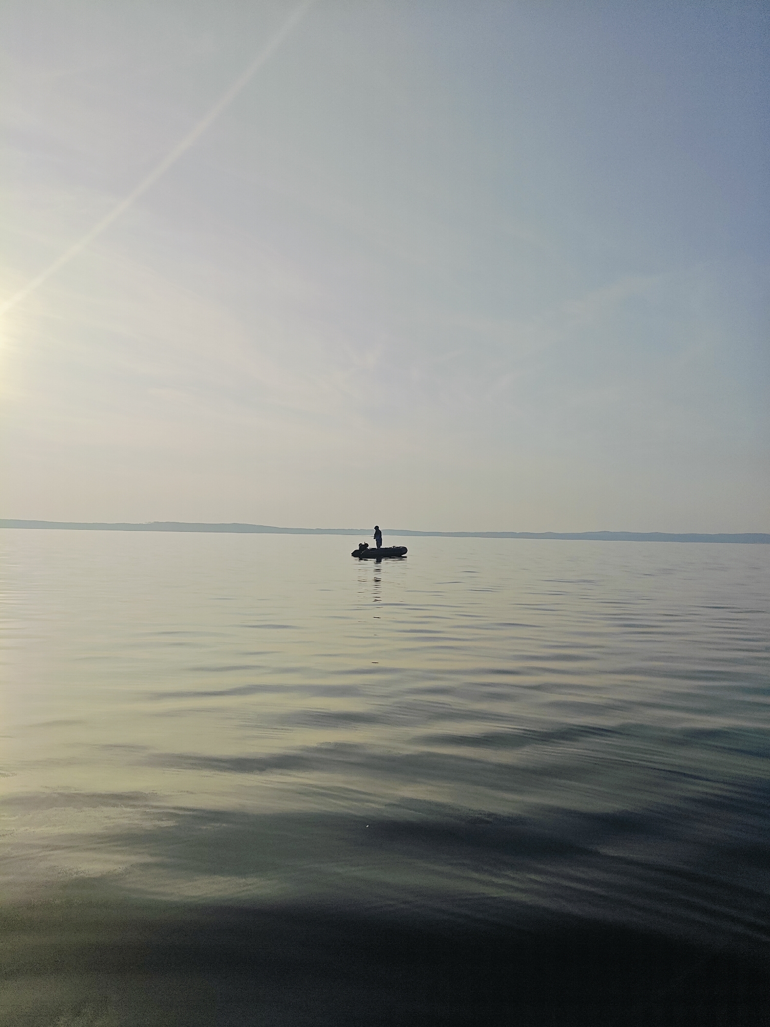 A person stands in a small inflatable boat on a calm body of water. The person is silhouetted against the bright, overcast sky and appears to be fishing. The water has gentle ripples and reflects the soft light of the sky, creating subtle patterns. In the distance, a faint, low-lying landmass forms a thin, dark line on the horizon. The overall mood of the image is peaceful and solitary, suggesting an early morning or late afternoon setting due to the diffused light. There is no visible text in the image. The scene is likely Chaykovsky, Russia, given the context.