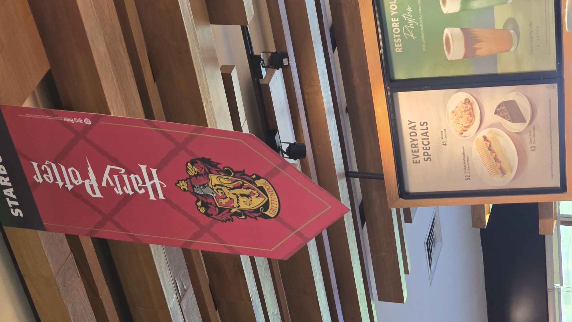 The image shows the interior of a Starbucks location. A Gryffindor house banner with the Harry Potter logo is displayed next to a menu board advertising "EVERYDAY SPECIALS" with items such as Almond Croissant, Smoked Beef Mushroom & Cheese Panini, and Classic Dark Chocolate Cake. The upper part of the board presents images of Vienna Creamy Latte and another drink with the caption, "RESTORE YOU Rhythm." The Starbucks appears to be located in Denpasar, Indonesia.