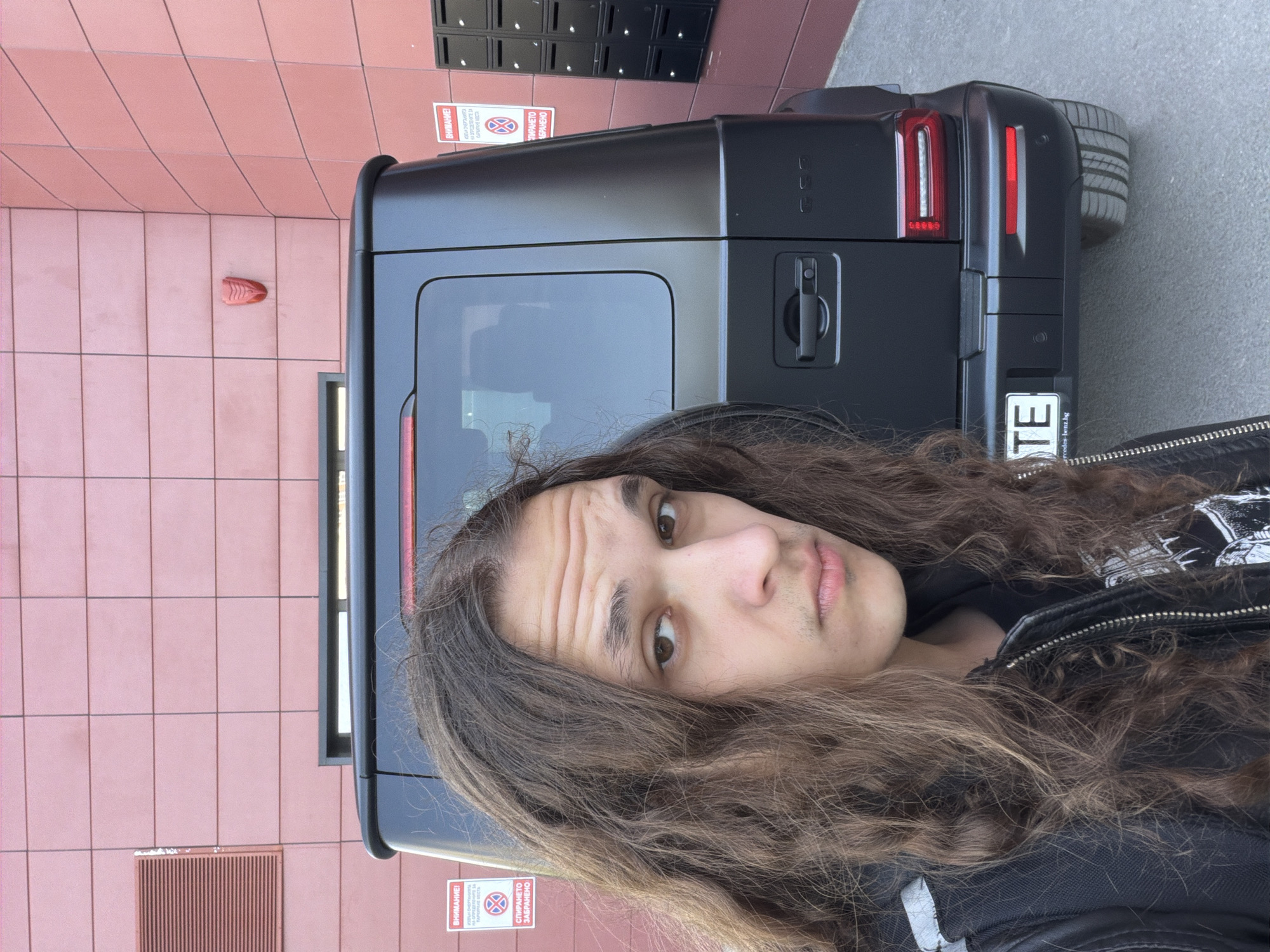 A person with long, wavy brown hair takes a selfie, holding the camera vertically, in an urban setting in Plovdiv, Bulgaria. The individual, wearing a black leather jacket over a dark shirt, looks directly at the camera with a neutral to slightly serious expression, featuring visible forehead wrinkles.
Behind the person, the rear of a matte black Mercedes-Benz G-Wagen is prominently featured, displaying its characteristic taillights, the exterior-mounted spare tire, and part of a license plate frame that reads "mercedes-benz.bg". The vehicle is positioned next to a building with a facade of reddish-brown square tiles. Above and to the left of the G-Wagen, a section of black mailboxes is mounted on the wall.
Two red warning signs are visible on the tiled wall. The first, partially obscured by the person's hair and the vehicle, displays "ВНИМАНИЕ!" (Attention!), "НА ОПРЕДЕЛЕНИТЕ ЗА" (For designated ones for), and "СПИРАНЕТО ЗАБРАНЕНО" (Stopping prohibited). A second, smaller sign, also says "ВНИМАНИЕ!" and "СПИРАНЕТО ЗАБРАНЕНО". Part of the vehicle's license plate shows the letters "TE". The scene is well-lit by daylight, suggesting clear weather.