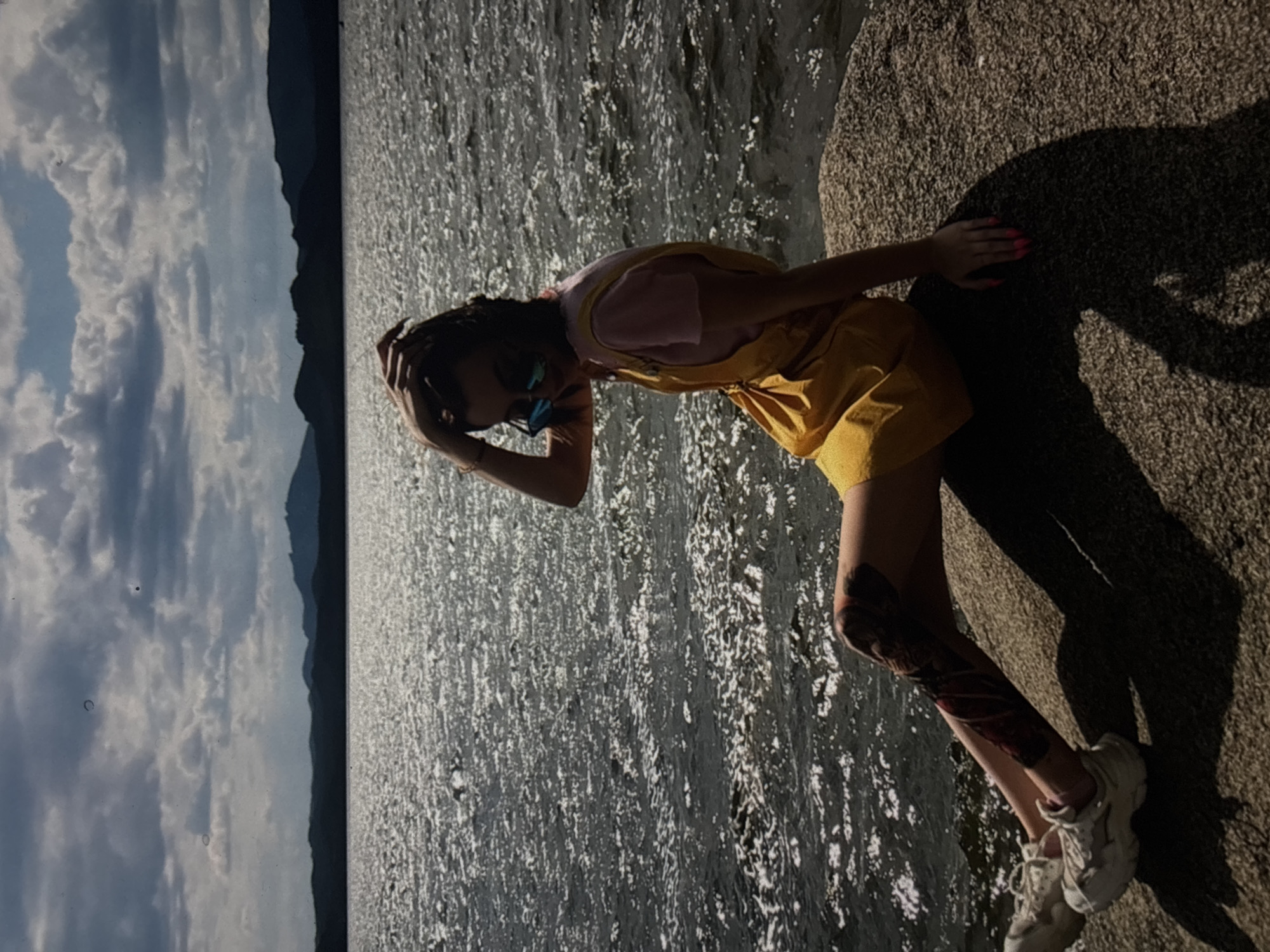 A young woman is sitting on a large rock by a body of water. She is wearing a yellow overall dress over a pink t-shirt and white sneakers. She has sunglasses on and her hand is resting on her head. Her legs are visible and she appears to have tattoos on her left leg. The water is choppy and reflecting the sunlight. In the background, there are dark hills or mountains under a cloudy sky. The scene is outdoors, likely in a natural setting, and the lighting suggests it is daytime, possibly late afternoon or early morning due to the strong shadows. The location is identified as the city of Omsk, Russia.