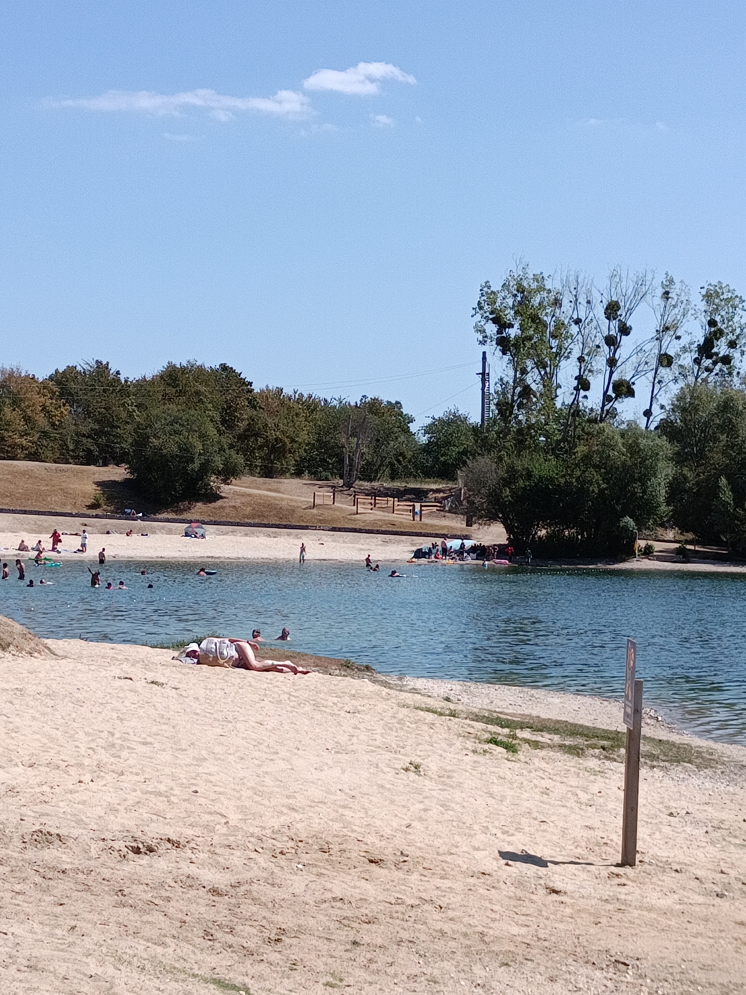 The image shows a sunny day at a sandy beach next to a body of water, likely a lake or reservoir, in the city of Le Mesnil-sous-Jumièges, France. The water is calm and blue, with several people swimming and wading in it. The beach is populated with sunbathers, some lying on towels and others sitting. In the background, there is a line of trees and vegetation, with a few buildings and structures visible in the distance. A wooden signpost stands on the beach in the foreground. The sky is a clear blue with a few scattered clouds.