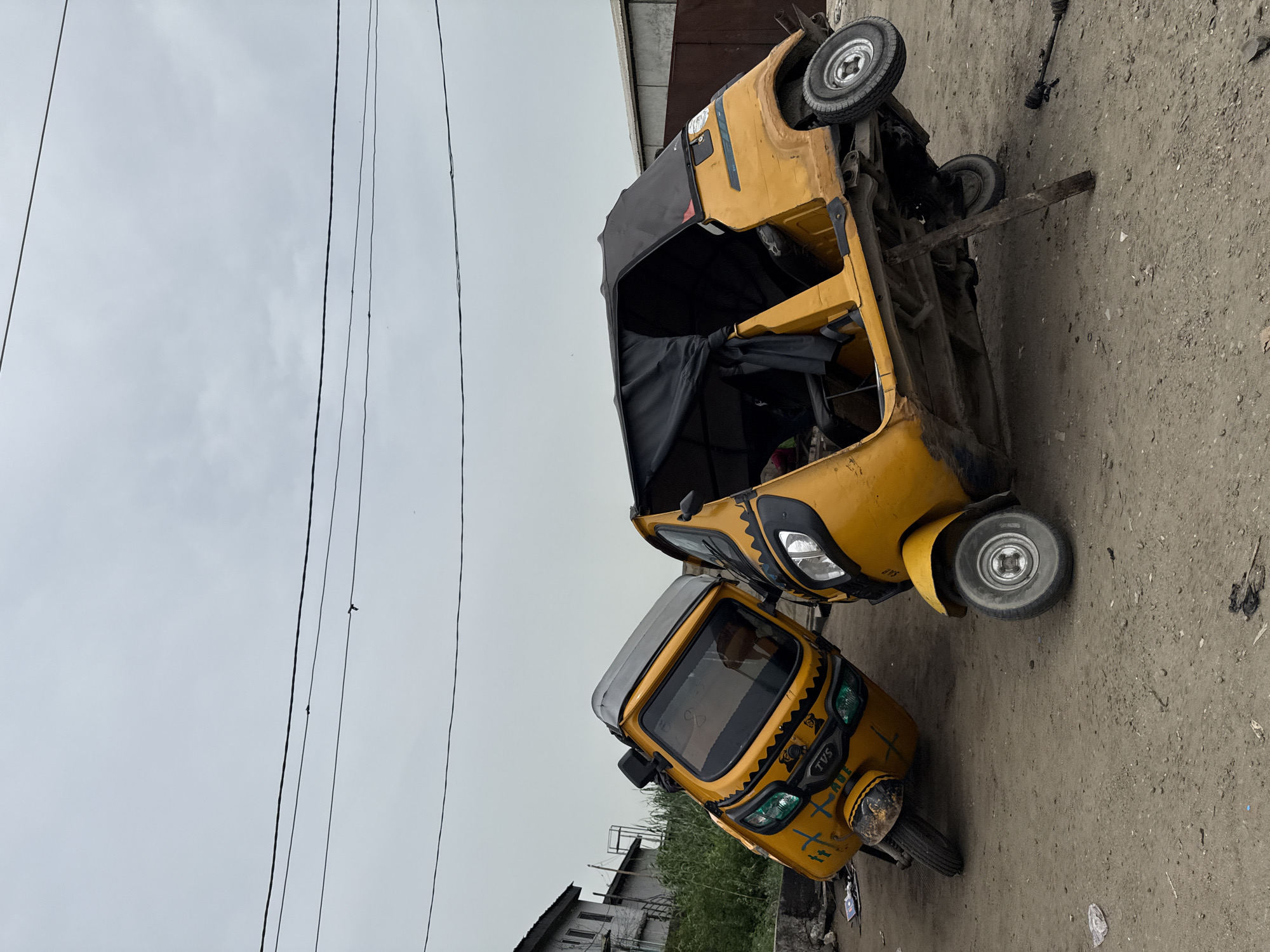 The image depicts two yellow-colored three-wheeled vehicles, commonly known as "keke" or "tuk-tuk", in Ado, Nigeria. One keke is upright and parked, while the other is overturned, suggesting possible damage or an accident. The environment shows a mix of urban elements with overhead electrical wires and a dirt/paved road. Vegetation and building structures are in the background. The sky is overcast. There are no visible people in the frame.