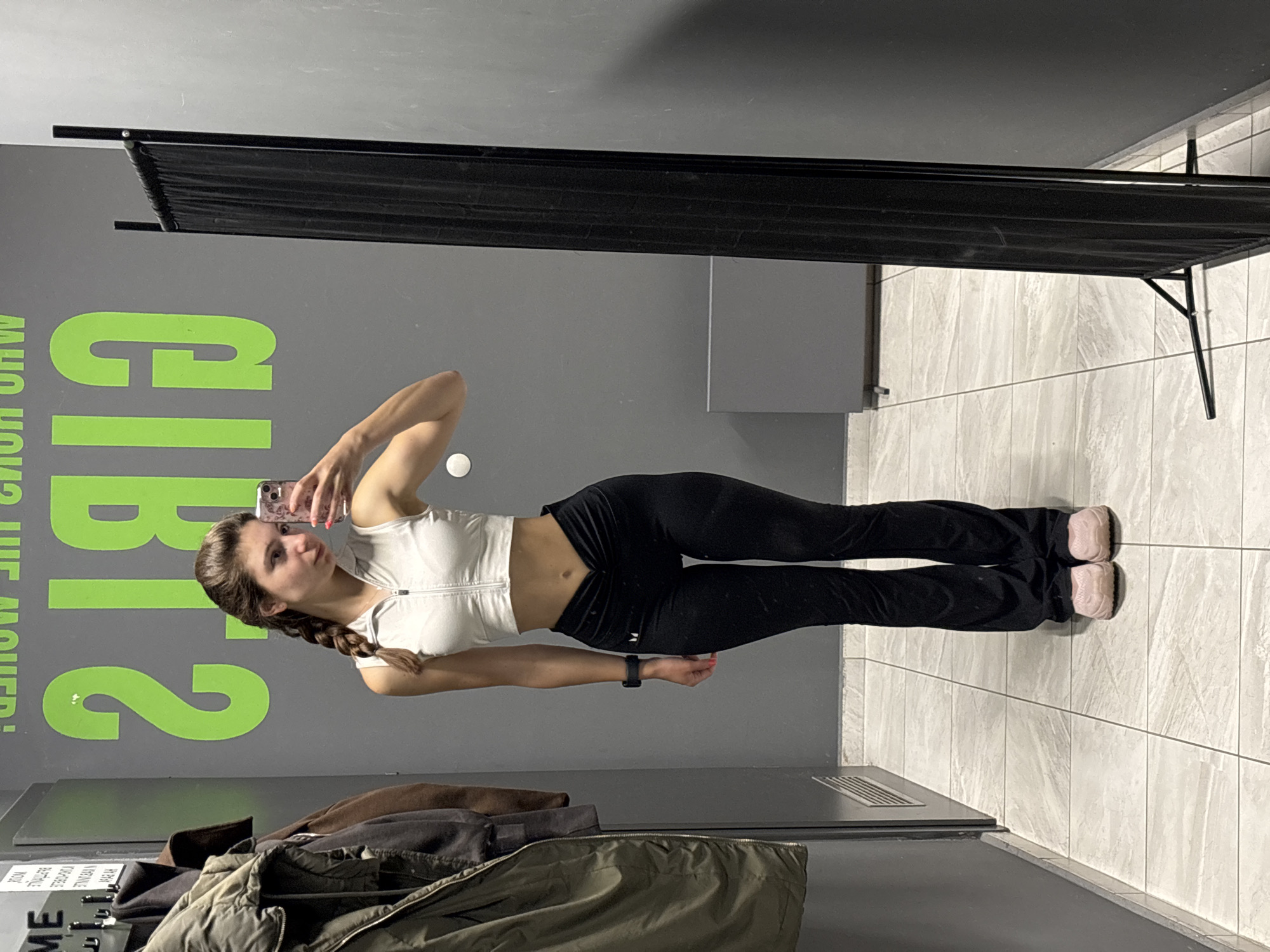 A young woman is taking a selfie in a gym changing room. She is wearing a white zip-up sports bra and black leggings. Her hair is in a braid. She is holding her phone in her right hand. There is a black folding screen behind her. The wall behind her has the words "Who's the mover?" written in green. There are clothes hanging on a rack in the bottom left corner of the image. The floor is tiled. It is impossible to confirm the location is Varna, Bulgaria.