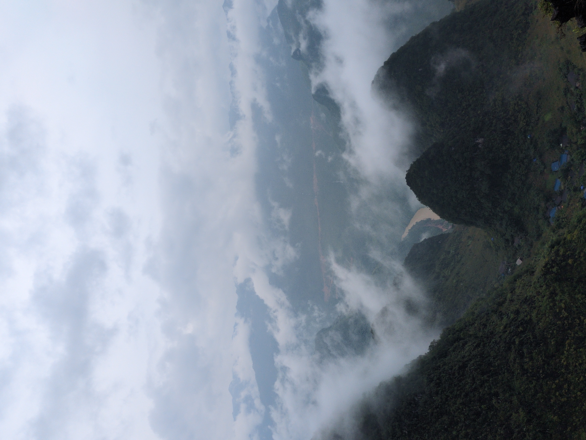 The image shows a mountainous landscape in Đồng Văn Commune, Vietnam, characterized by lush green peaks partially obscured by rolling clouds and mist. A winding river, appearing brownish in color, cuts through a valley between the mountains. In the foreground, the steep, verdant slopes of several mountains are visible, with some hints of small structures or dwellings nestled at their bases. The sky is overcast, contributing to the overall misty and dramatic atmosphere of the scene.